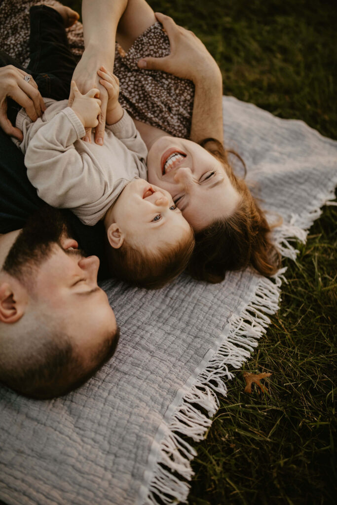 a family (father, mother, son) laying on a picnic blanket, cuddling together and laughing during a family session