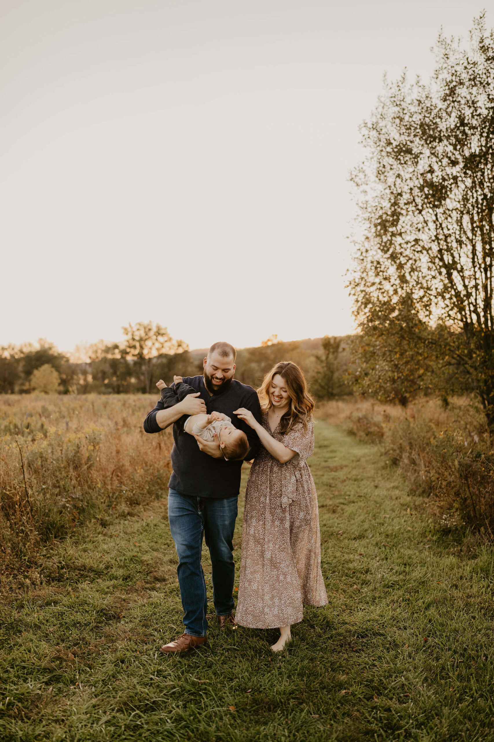 a family (mother, father, and son) walking toward the camera through a field at sunset at Peace Valley Park. The son is being held upside down and giggling.