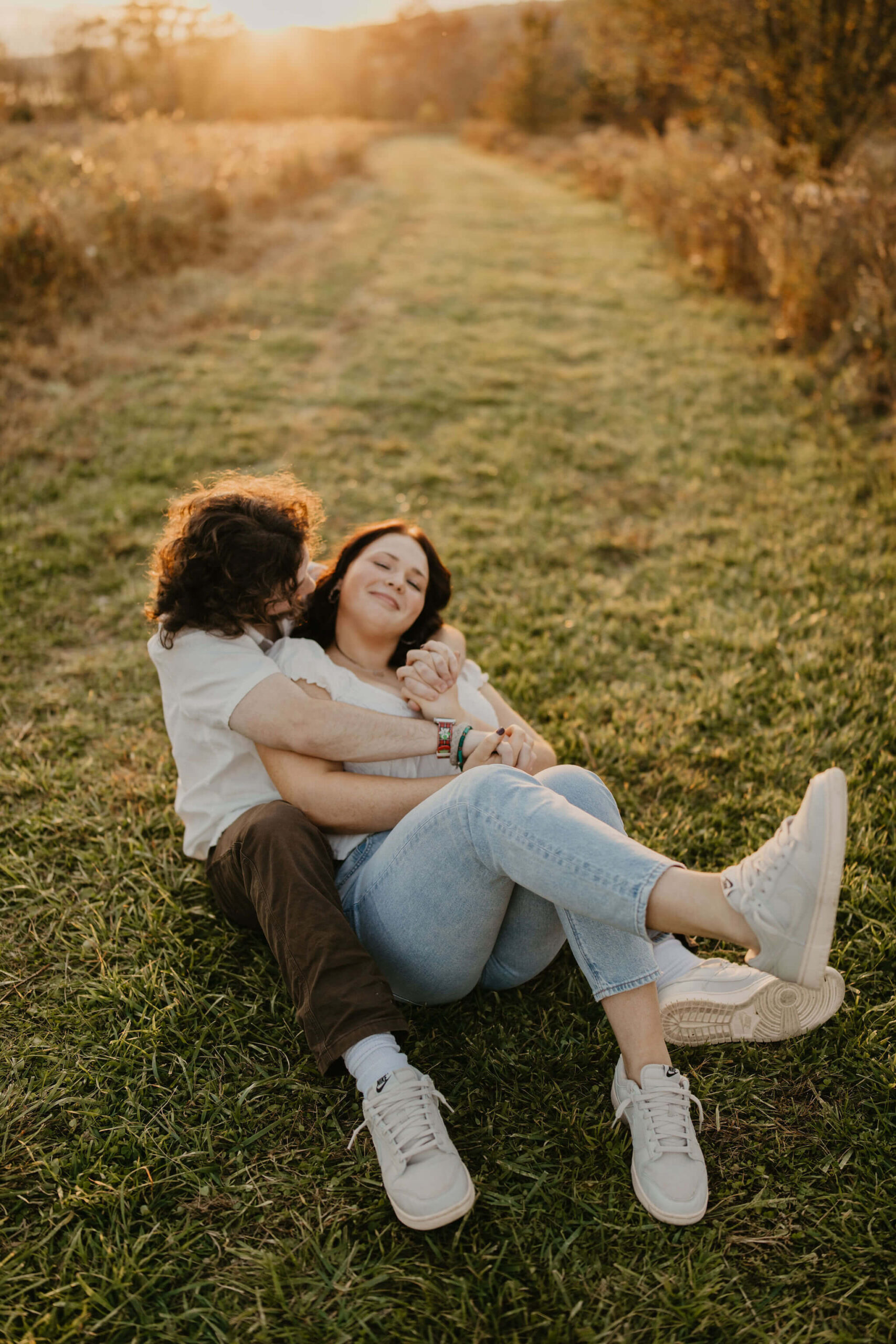 a couple cuddling in a field during golden hour. his arms are wrapped around her and she is smiling softly at the camera during a couples session