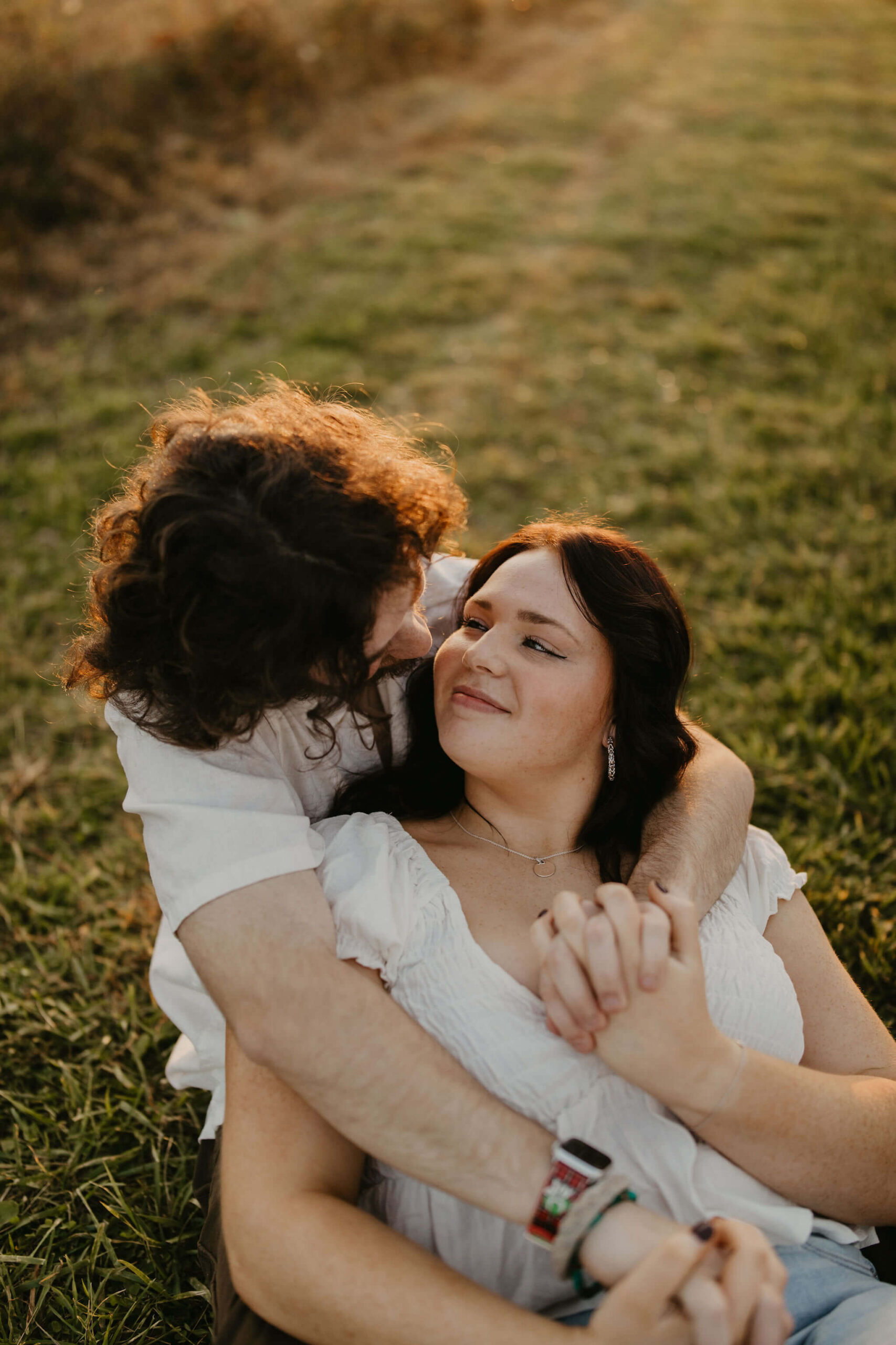 a couple sitting in a field cuddling. his arms are wrapped around her and they are smiling at each other during their couples session