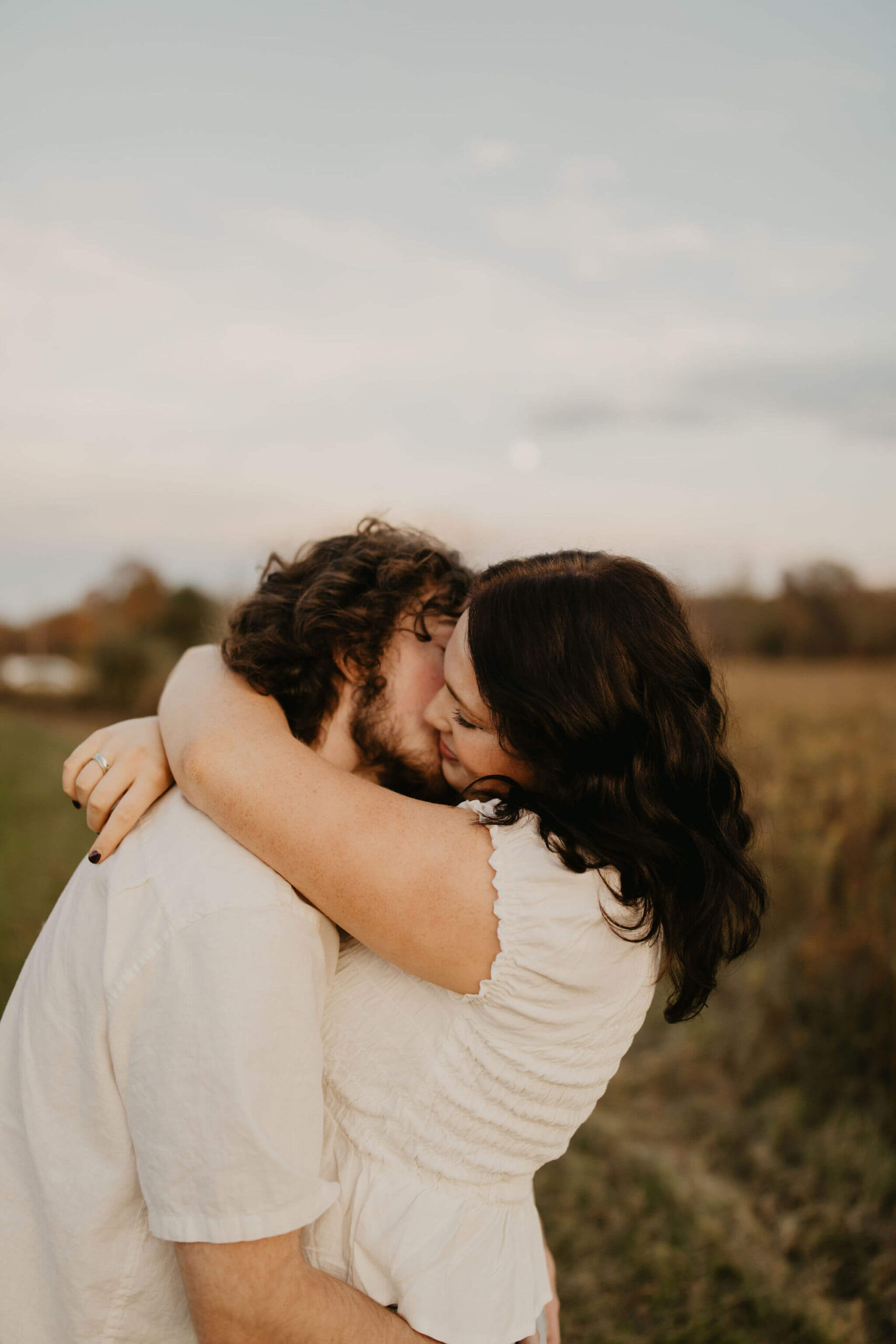 a boy kissing his girlfriend on the cheek in a field at sunset. the moon is softly rising behind them in doylestown pa