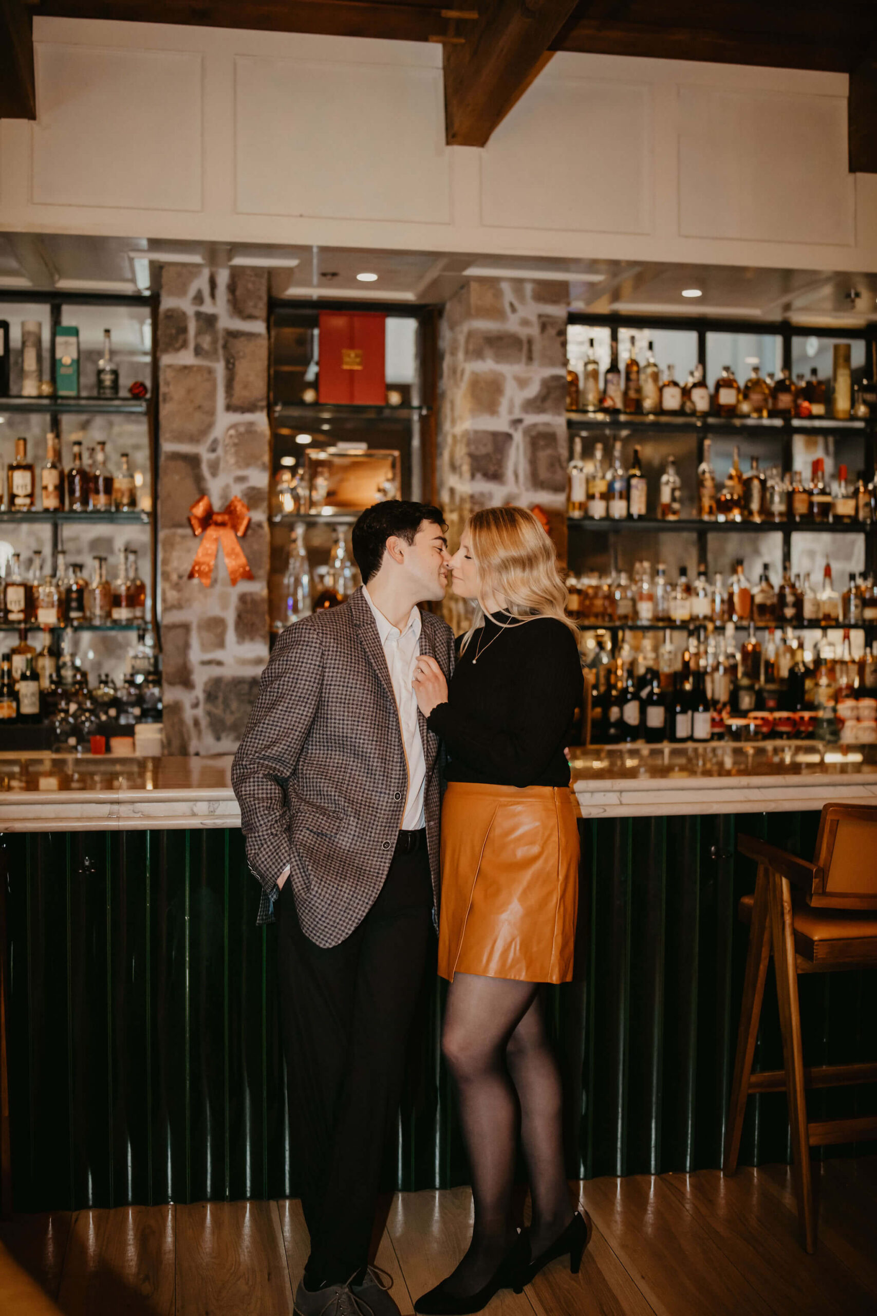 man and woman standing against a bar with a full liquor shelf behind it, she is holding the lapel of his suit and bringing him in for a kiss during their engagement session