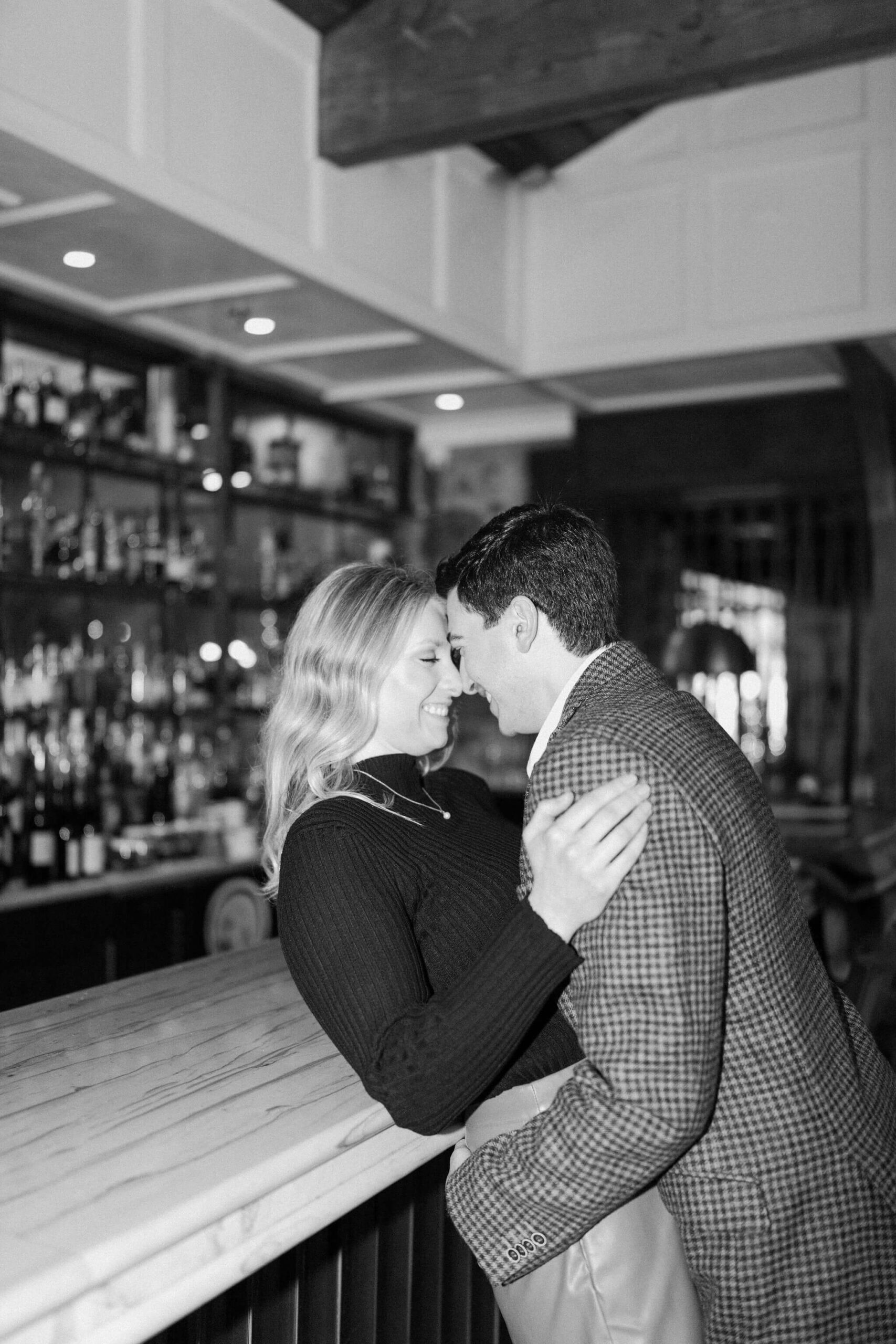black and white image of a couple at a bar, foreheads leaning together and smiling, with the man leaning the woman back over the bar during their engagement session