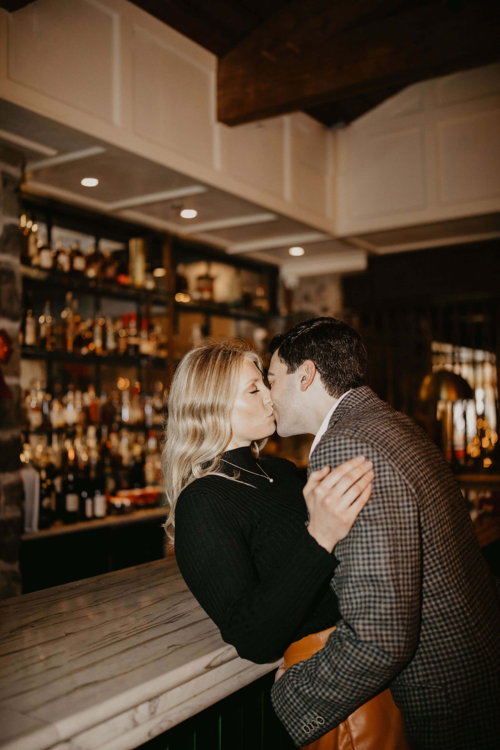 close up image of a man kissing a woman with her back pressed against a bar in New Hope during their engagement session, a full liquor shelf visible behind them