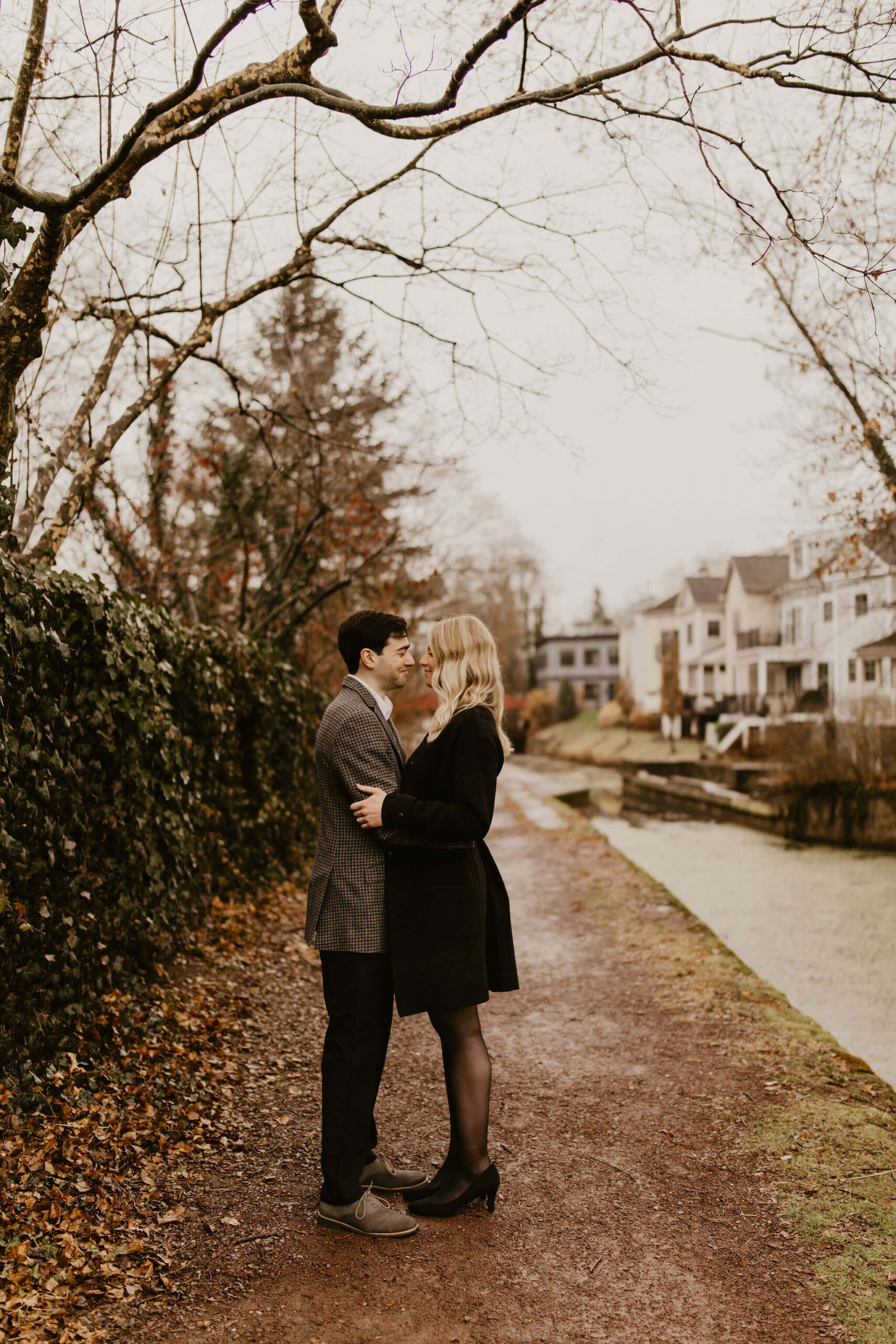 man (brown hair, brown checkered suit) and woman (long blonde hair, long black jacket and tights) embracing next to a canal in New Hope and smiling at each other