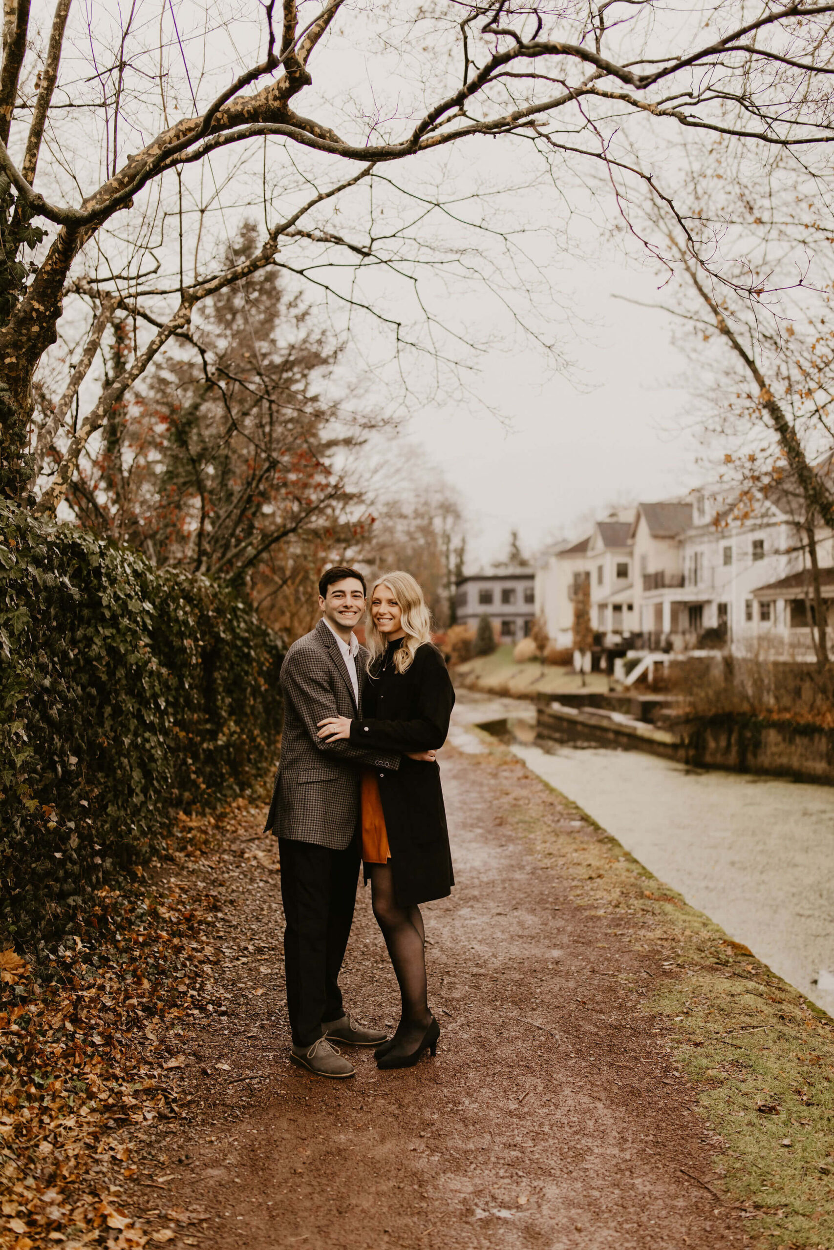 man (brown hair, brown checkered suit) and woman (long blonde hair, black shirt and brown skirt) holding each other on a walking trail next to a river, looking at the camera and smiling during their engagement session in New Hope
