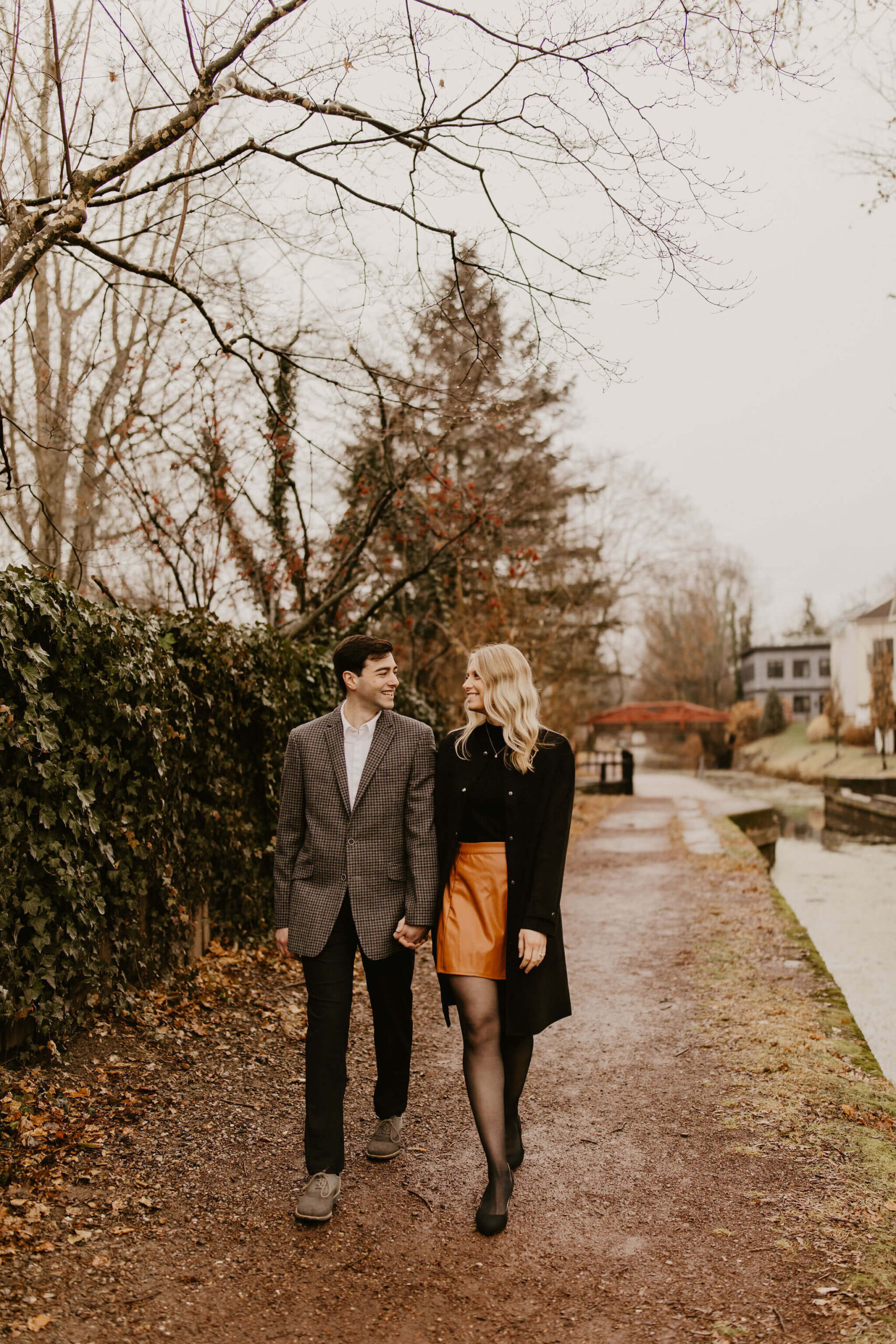 man (brown hair, brown checkered suit) and woman (long blonde hair, black shirt and brown skirt) walking toward the camera along a path and smiling at each other during their engagement session