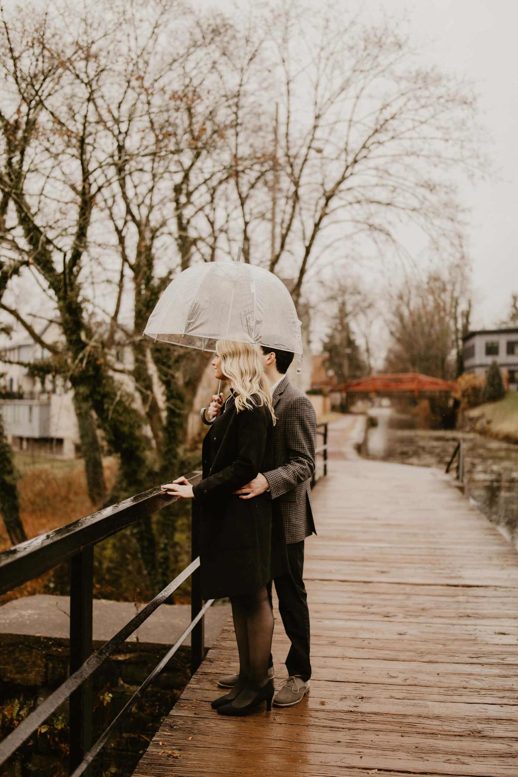man with his arm around his fiance's back, looking off camera to the left while standing on a bridge in New Hope, holding a clear umbrella over them both during their engagement session