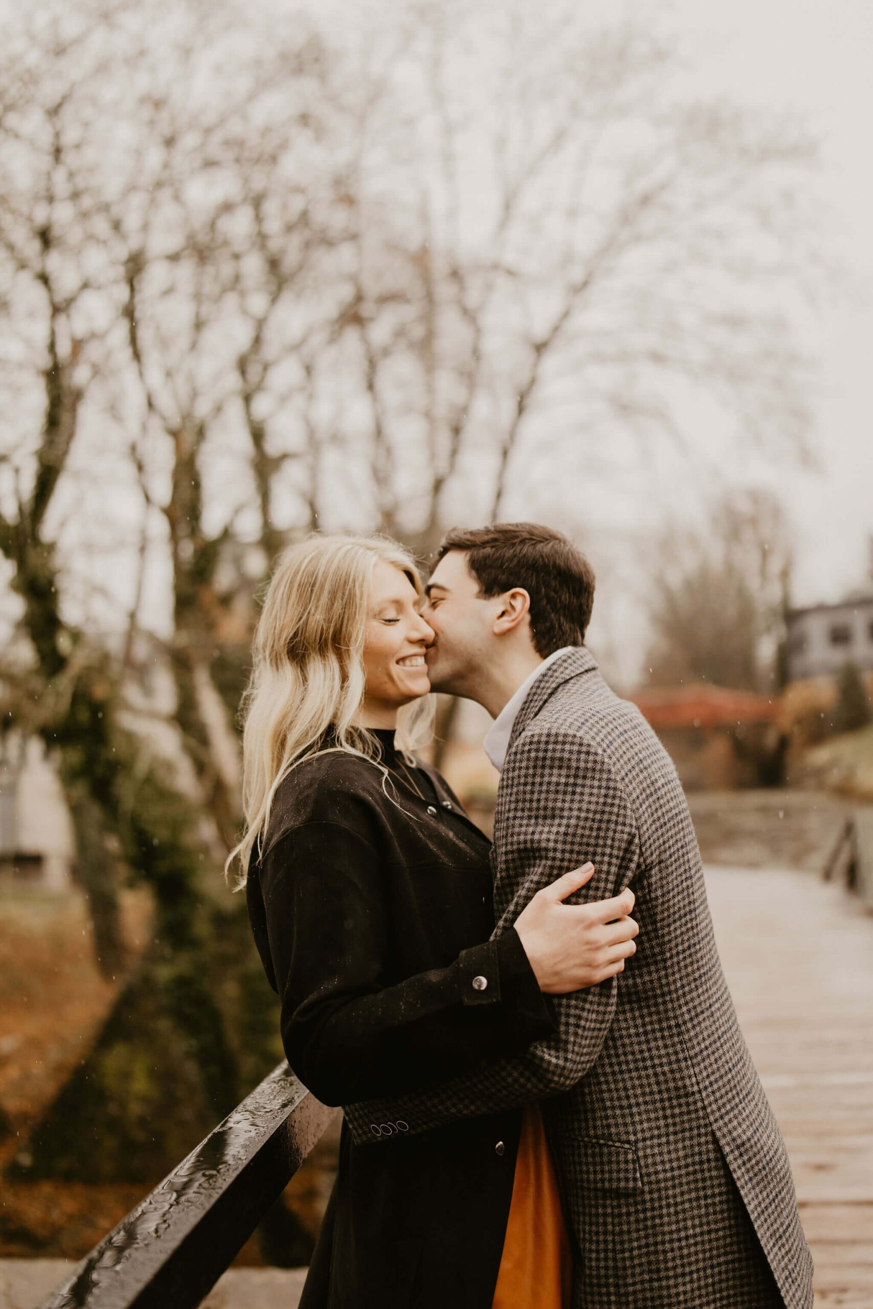 man kissing a woman on the cheek on a walking bridge, both smiling, during their engagement photo session in Bucks County, Pennsylvania