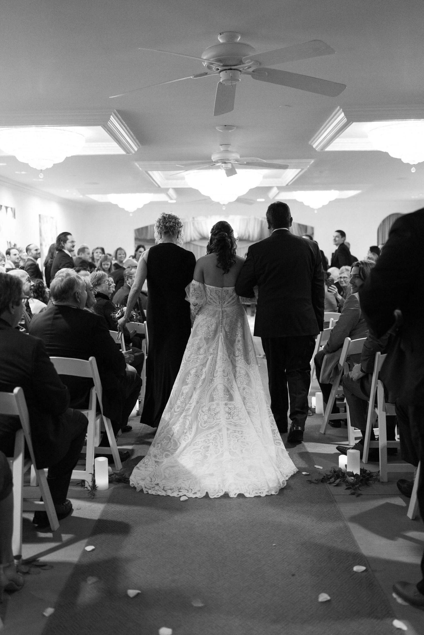 black and white image of a bride at her wedding being walked down the aisle by her mom and dad, on either side of her in lambertville new jersey