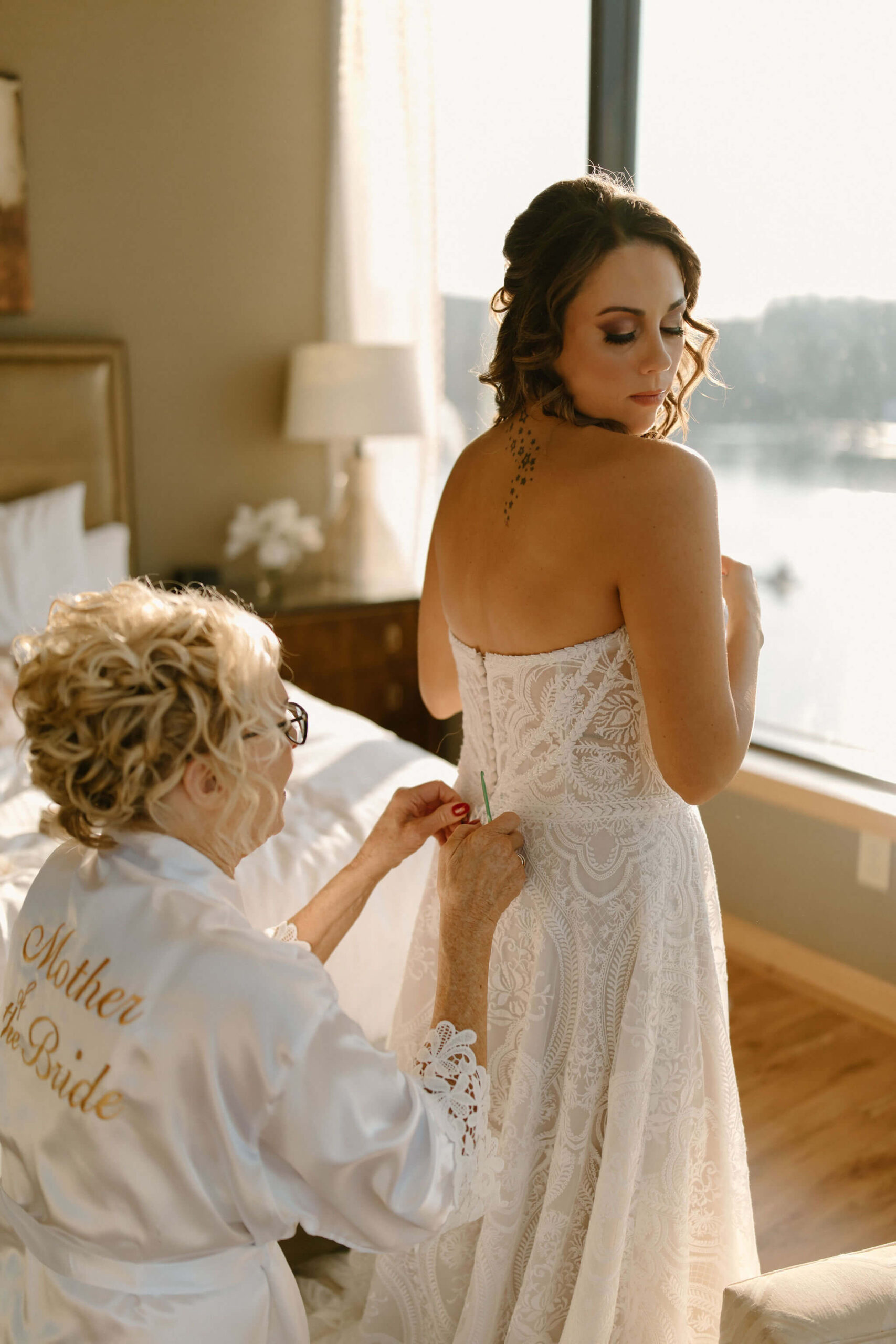 the mother of the bride in a white robe helping her daughter (the bride) button her wedding dress with a crochet hook in lambertville new jersey