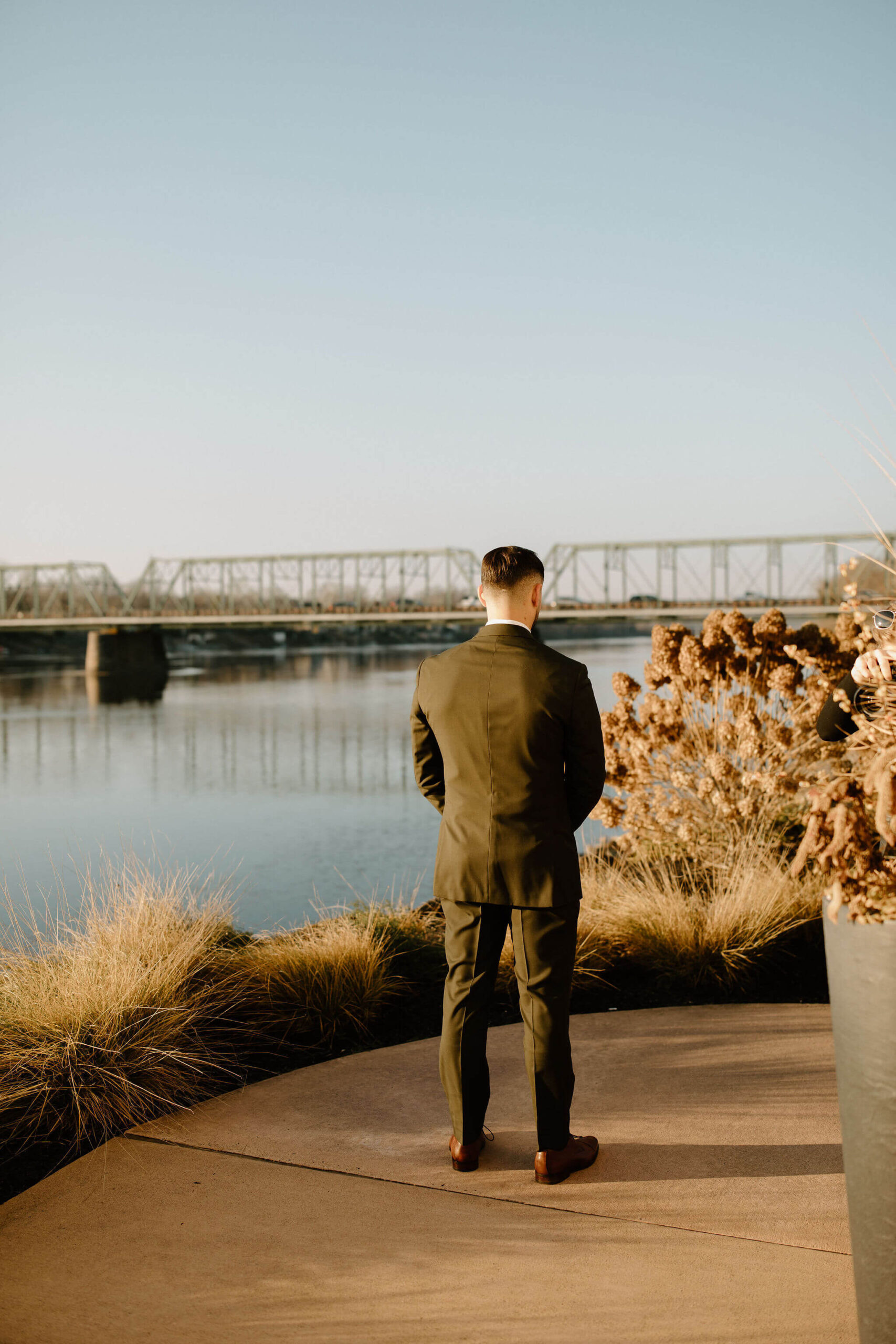 male groom (brown hair, green suit) facing away from the camera toward a walking bridge, waiting for the first look with his bride, the lambertville new jersey bridge in the background