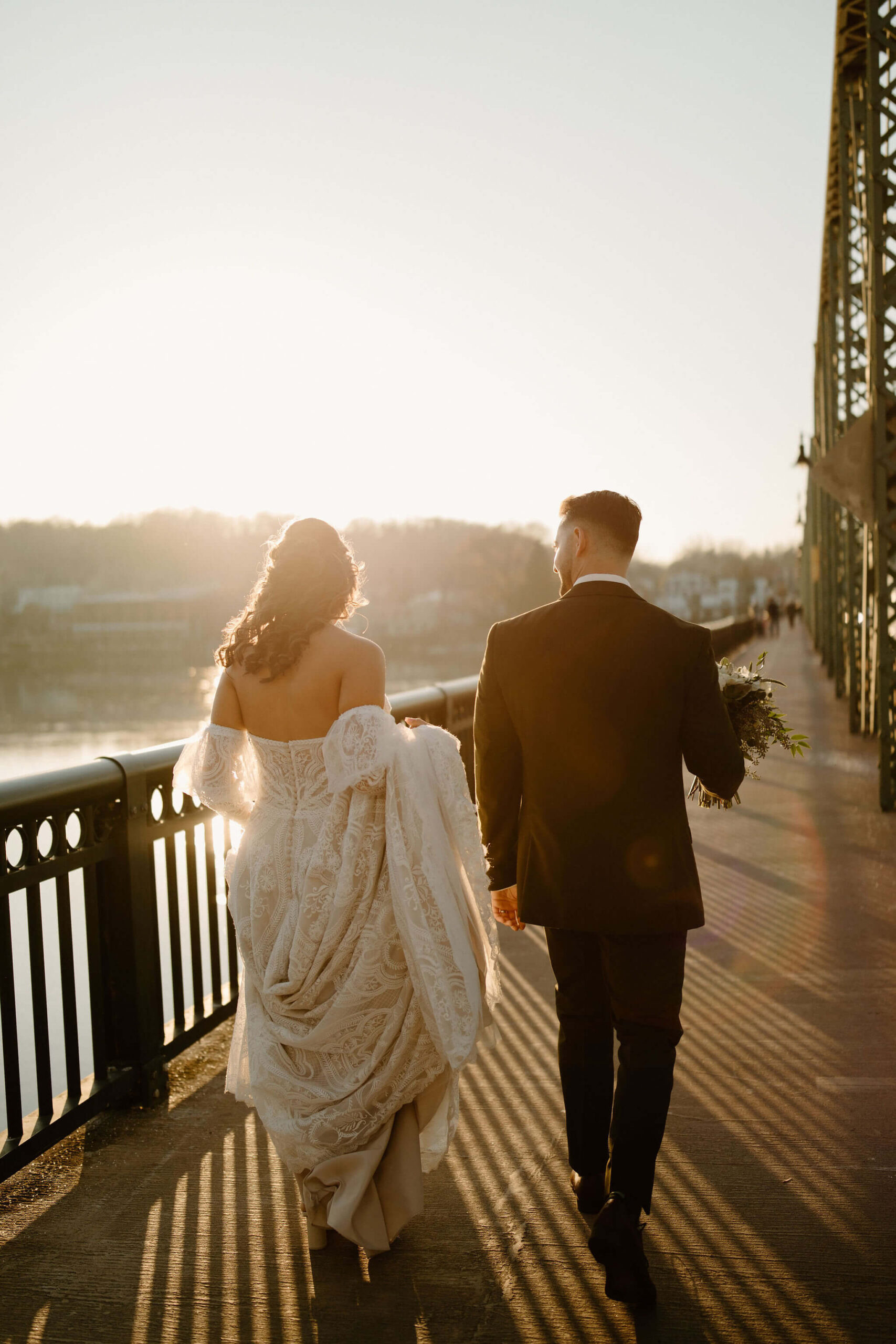 bride and groom walking on a bridge across a river, toward the sunset away from the camera, taking their wedding pictures in New Hope, Pennsylvania