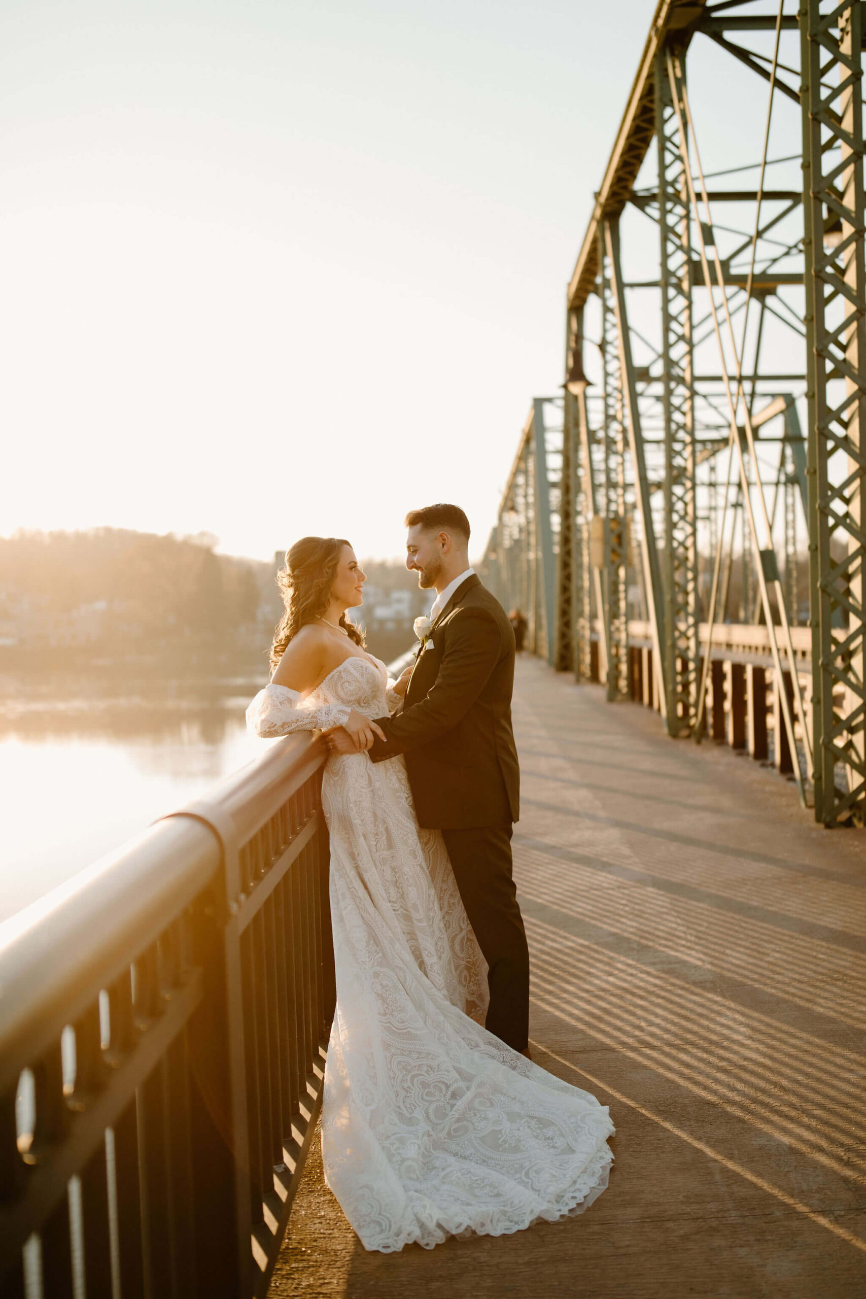 Bride (off-white boho dress) with her back against a railing of a bridge overlooking a river at sunset. The Groom has his arms around her waist and they are smiling at each other at their new jersey wedding