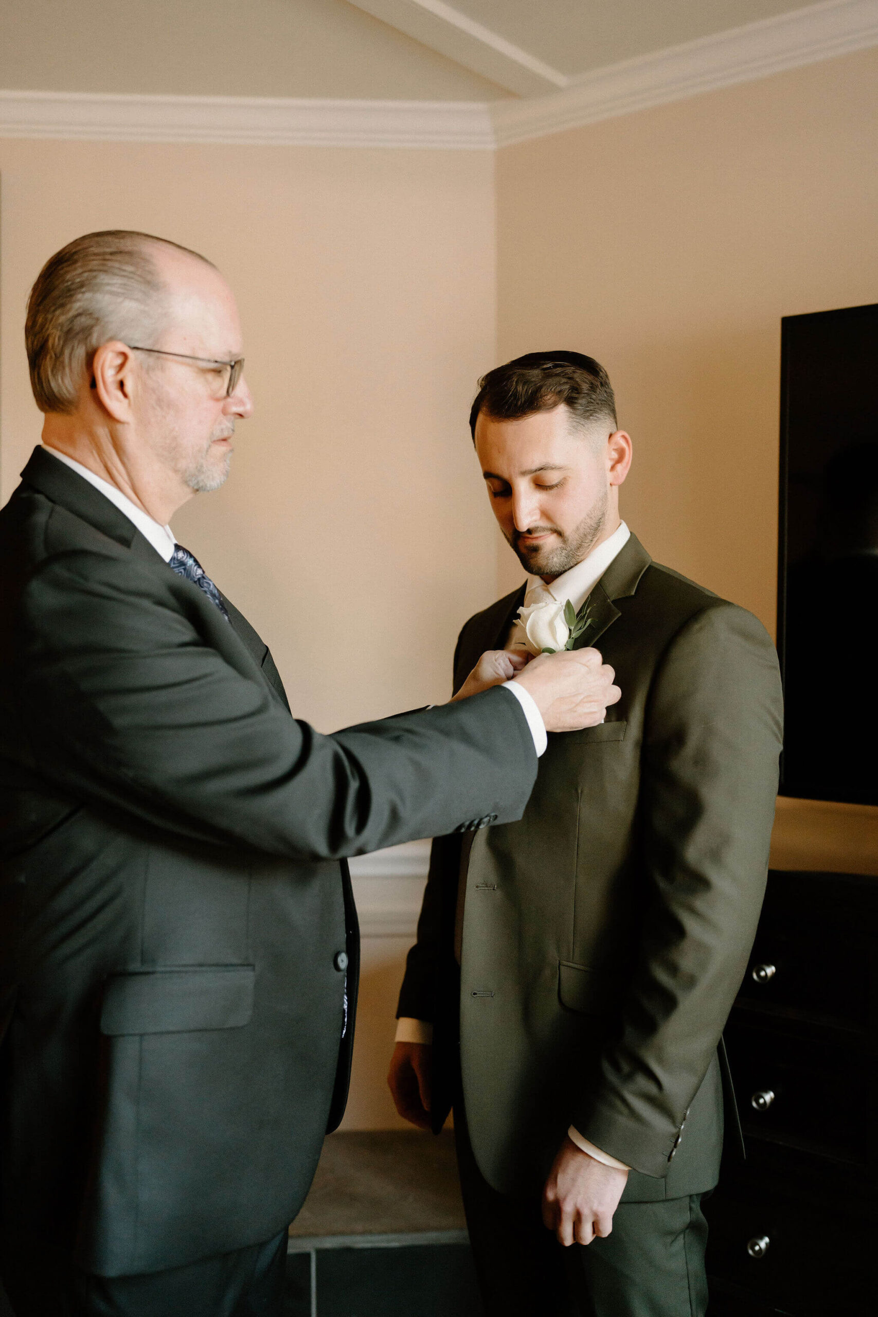 father of the groom (white slicked back hair and a black suit) helping the groom (brown hair and green suit) to pin his bouttoniere, at a wedding venue in lambertville new jersey