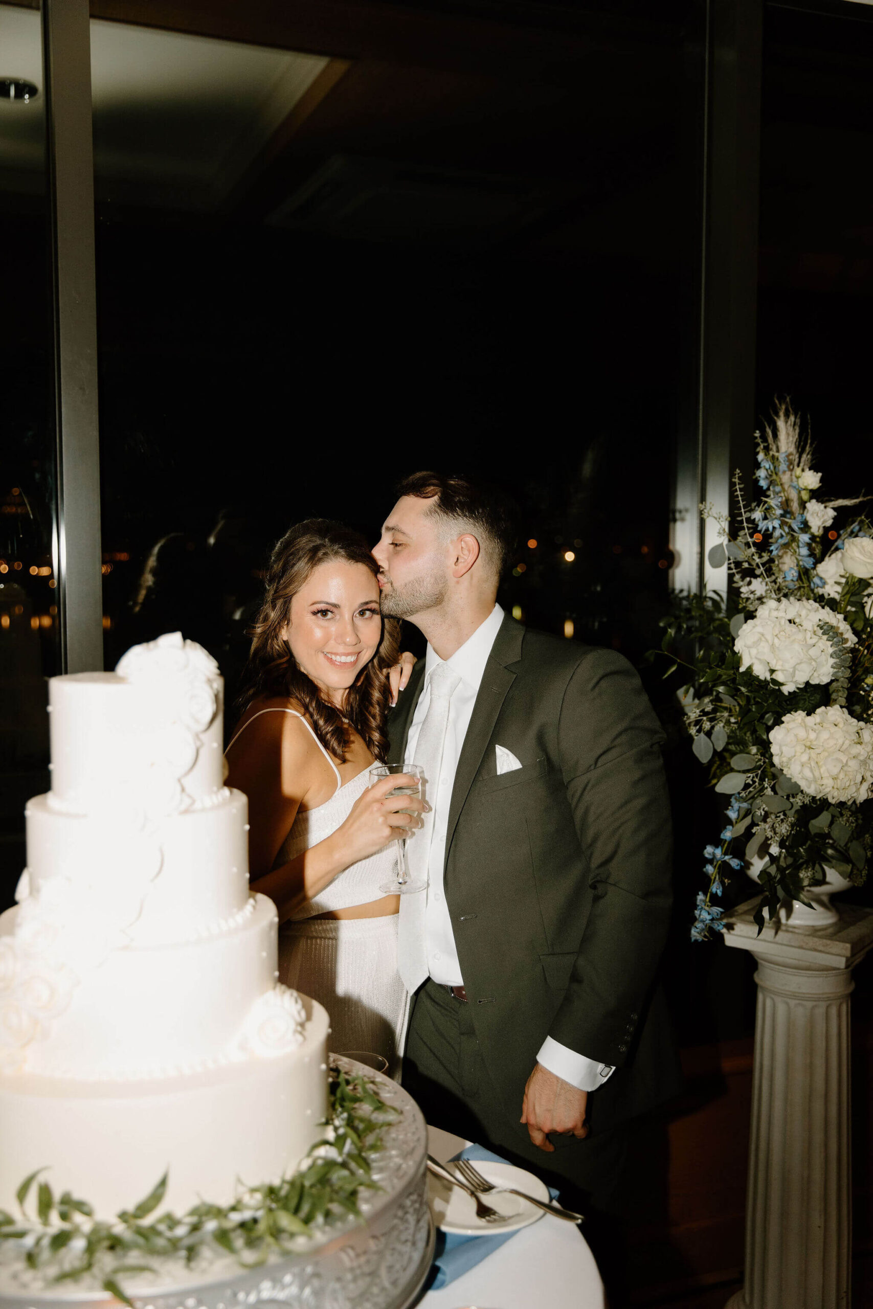 groom kissing bride on the cheek while bride smiles at the camera as they prepare to cut their wedding cake in lambertville new jersey