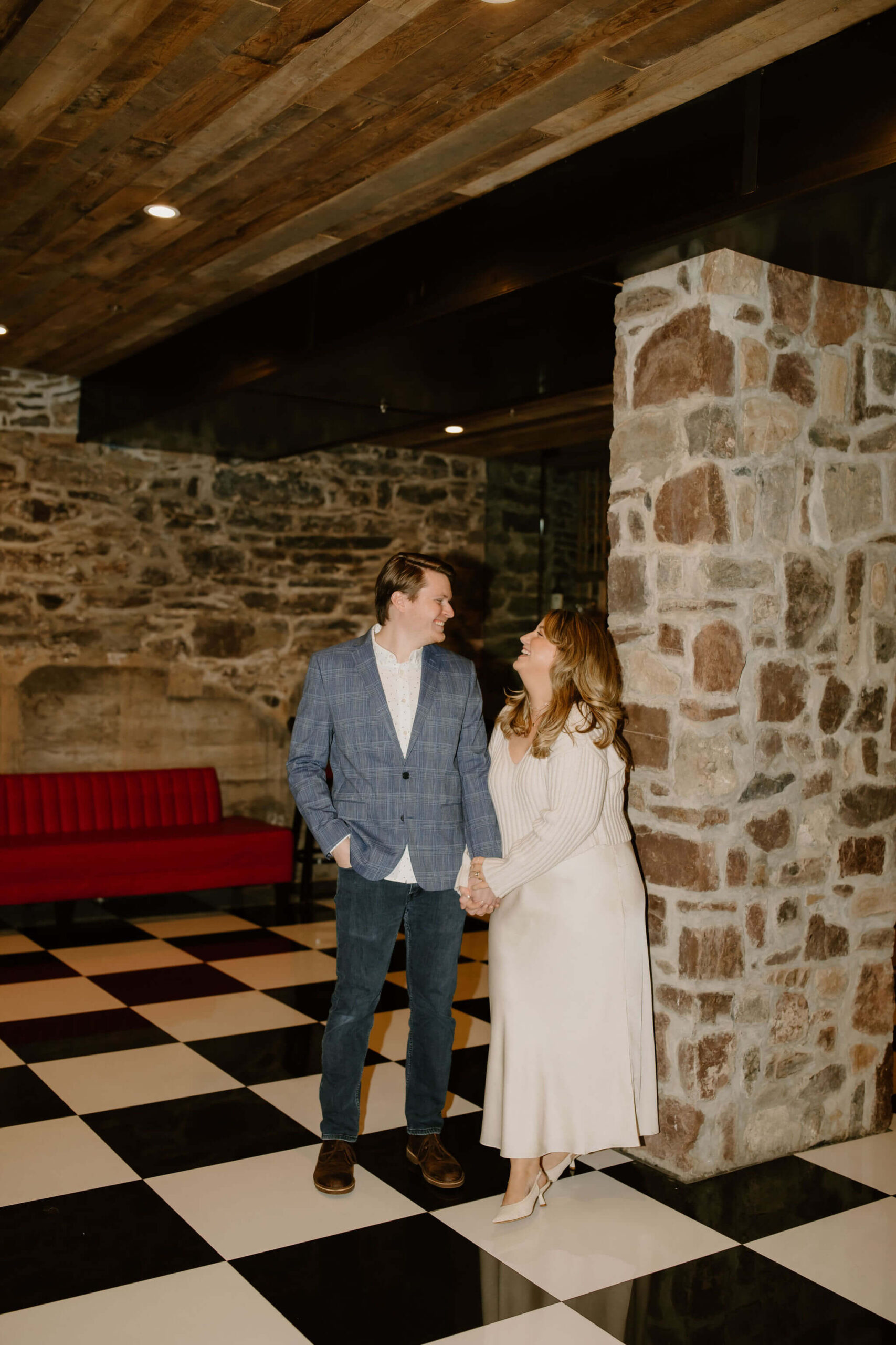 man (light brown hair, blue plaid suit jacket, jeans, and boots) holding hands with a woman (long blonde hair, white sweater, white skirt), looking at each other and smilng against a stone wall and black and white checkered floor for engagement photos
