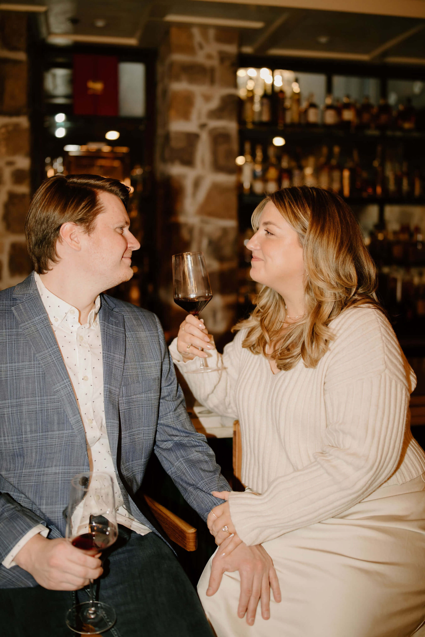 man and a woman seated at a bar, both holding wine glasses, with his hand on her knee, smiling softly at each other at the Logan Inn