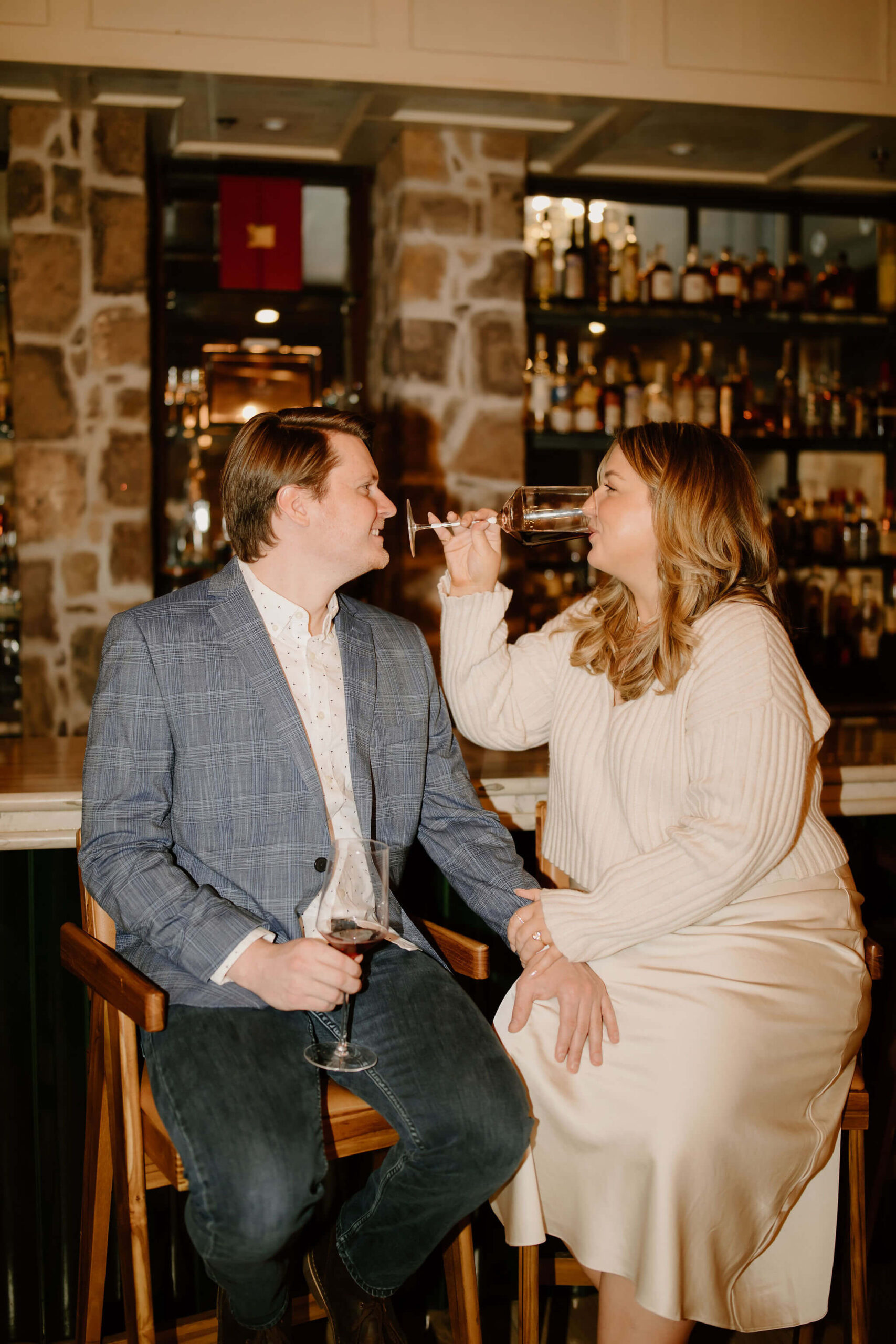 man and a woman seated at a bar, both holding wine glasses, with his hand on her knee, smiling softly at each other while she takes a sip of wine at the Logan Inn