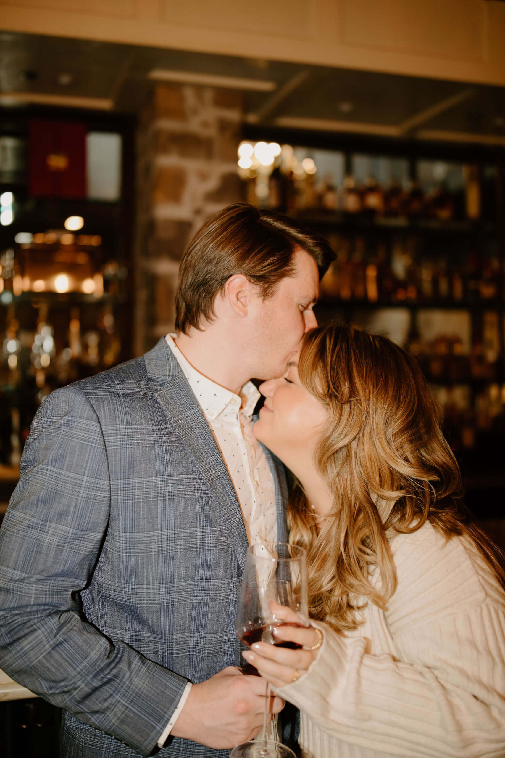 man standing and a woman seated at a bar at the Logan Inn, both holding wine glasses, while he kisses her on the forehead