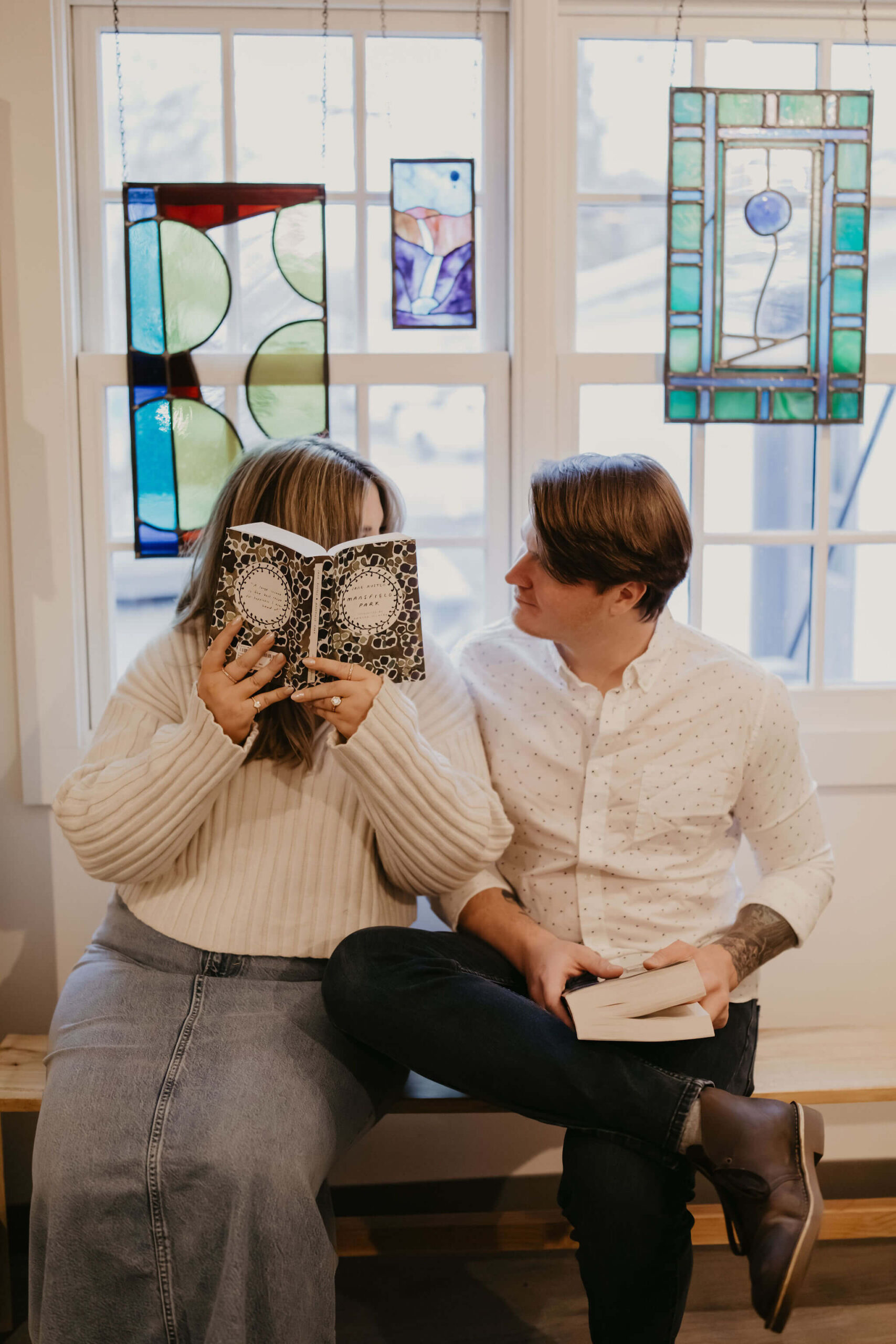 a couple sitting on a bench against a window, her holding a book open covering her face, while he holds a book in his lap looking at her during their engagement session
