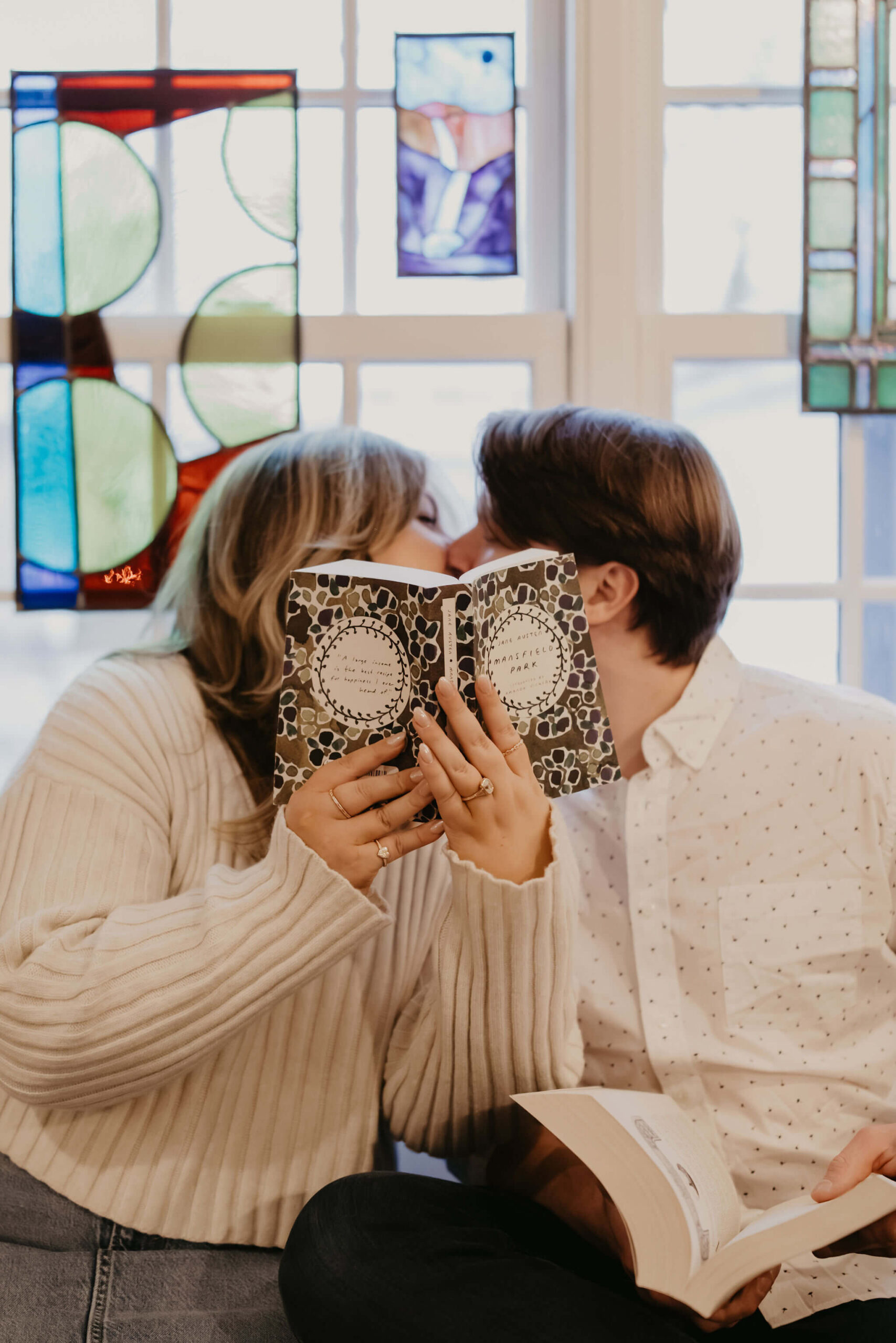 a couple sitting against a stained glass window, kissing behind an opened book during their engagement session