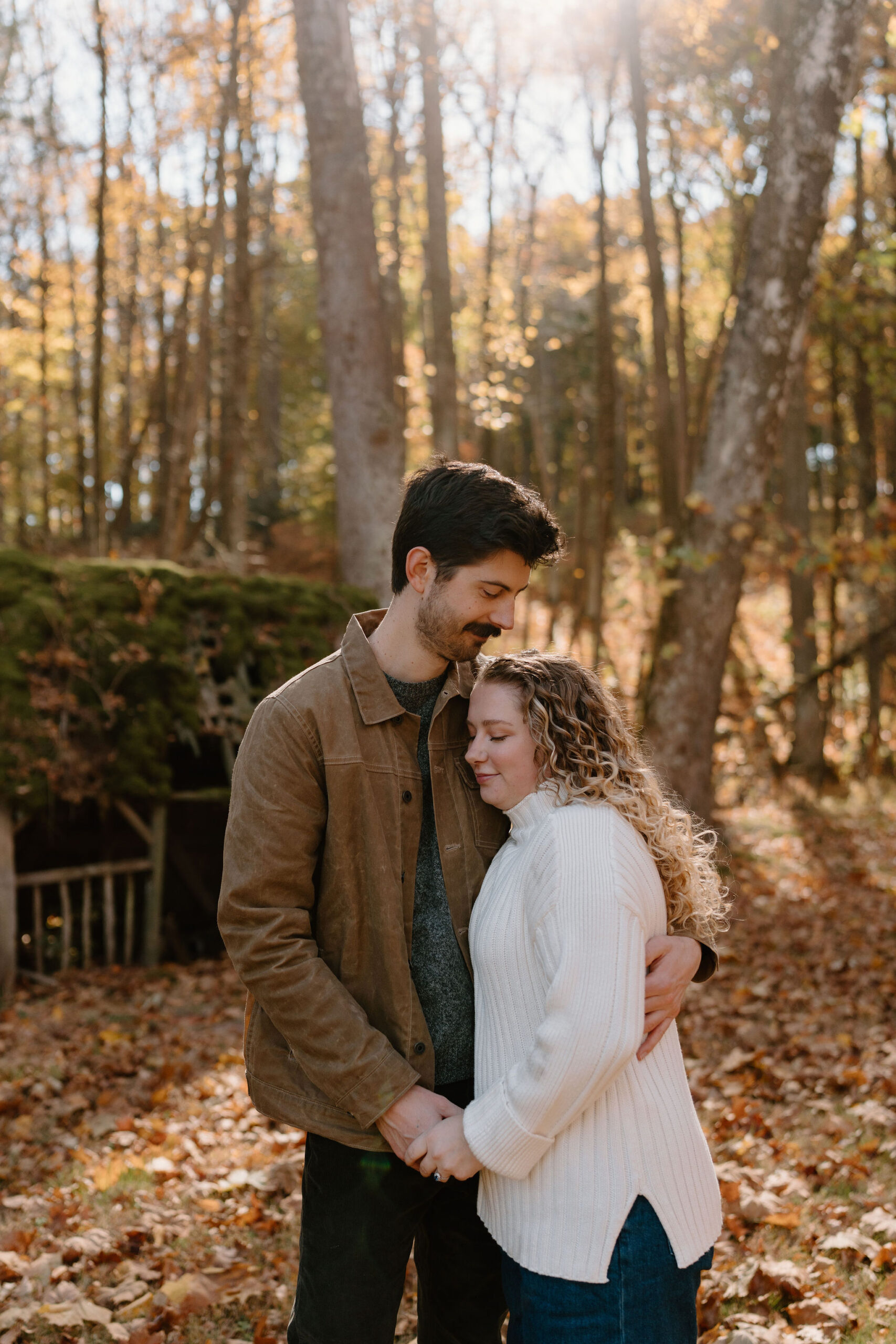 engagement photos of a couple in the woods on a sunny fall day, holding hands and leaning into each other.