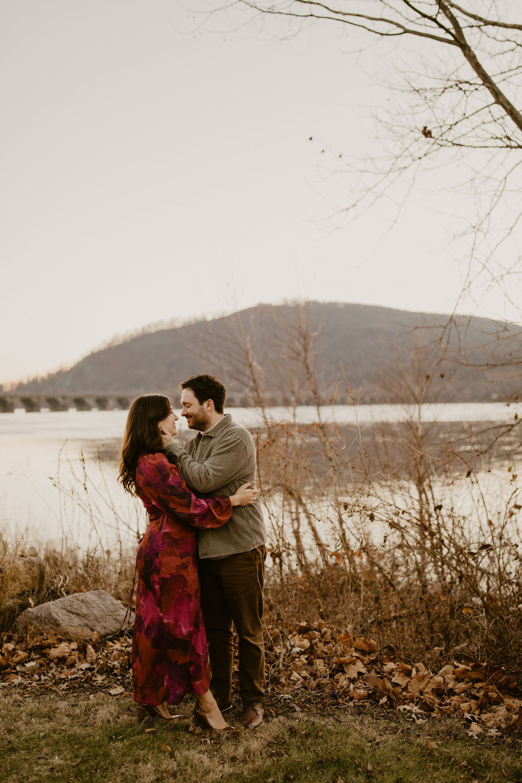 engagement photos of a couple embracing, about to kiss, in front of a river with a large bride going across it