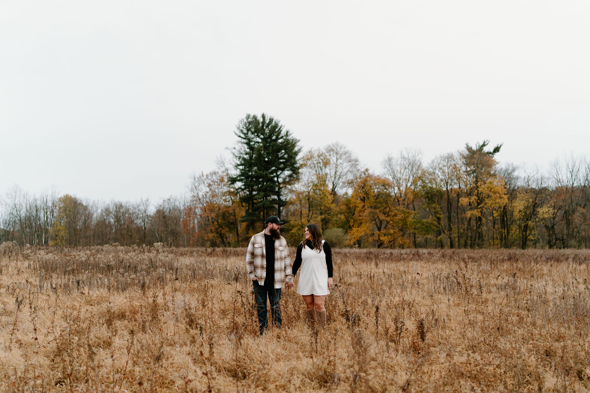 engagement session of a man with a beard, ball cap, and brown and white flannel, holding hands with his brunette fiance in a white jumper and black turtleneck, smiling at each other