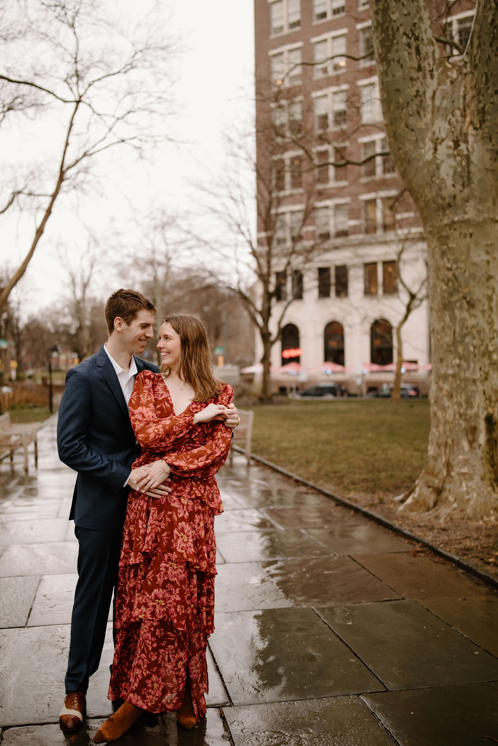 engagement session of a man in a navy blue suit and brown leather dress shoes, with his arms around his fiance in a red tiered flowy dress, smiling at each other in the city