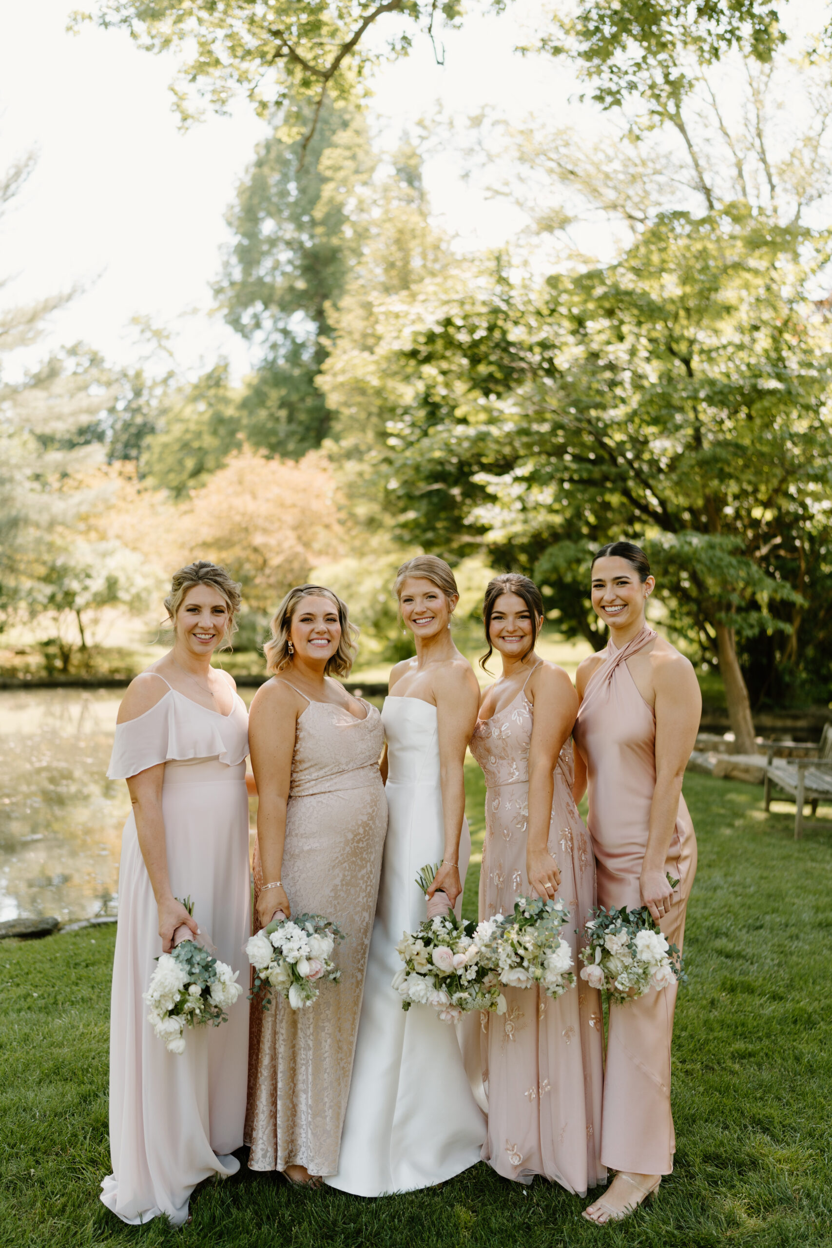 a bride (white dress) and four bridesmaids (light pink non-traditional mismatched dresses) holding flowers at their sides and smiling at the camera