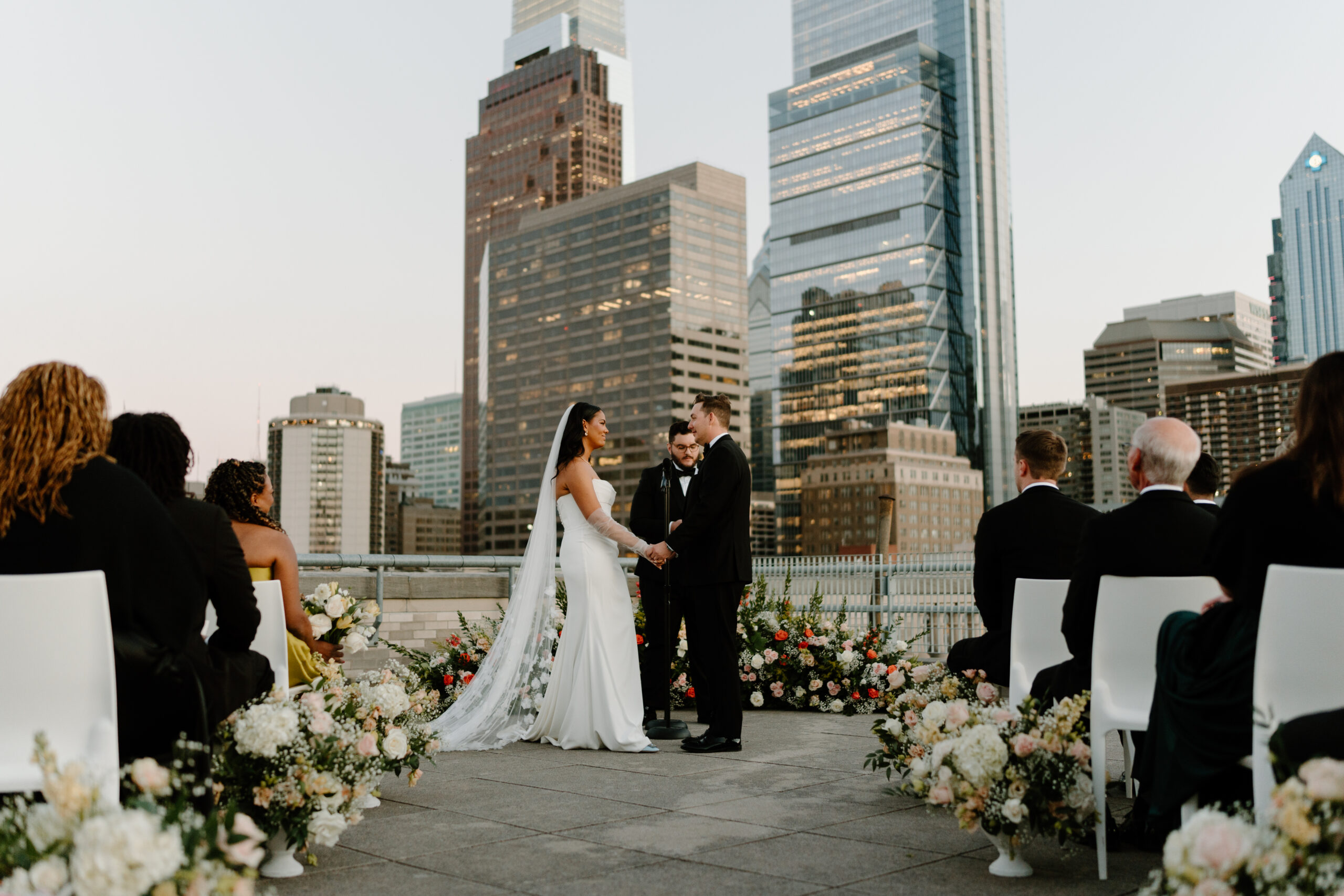 photograph of bride and groom holding hands and smiling softly at each other during their rooftop wedding ceremony, with guests sitting in white chairs watching, with the philadelphia skyling behind them