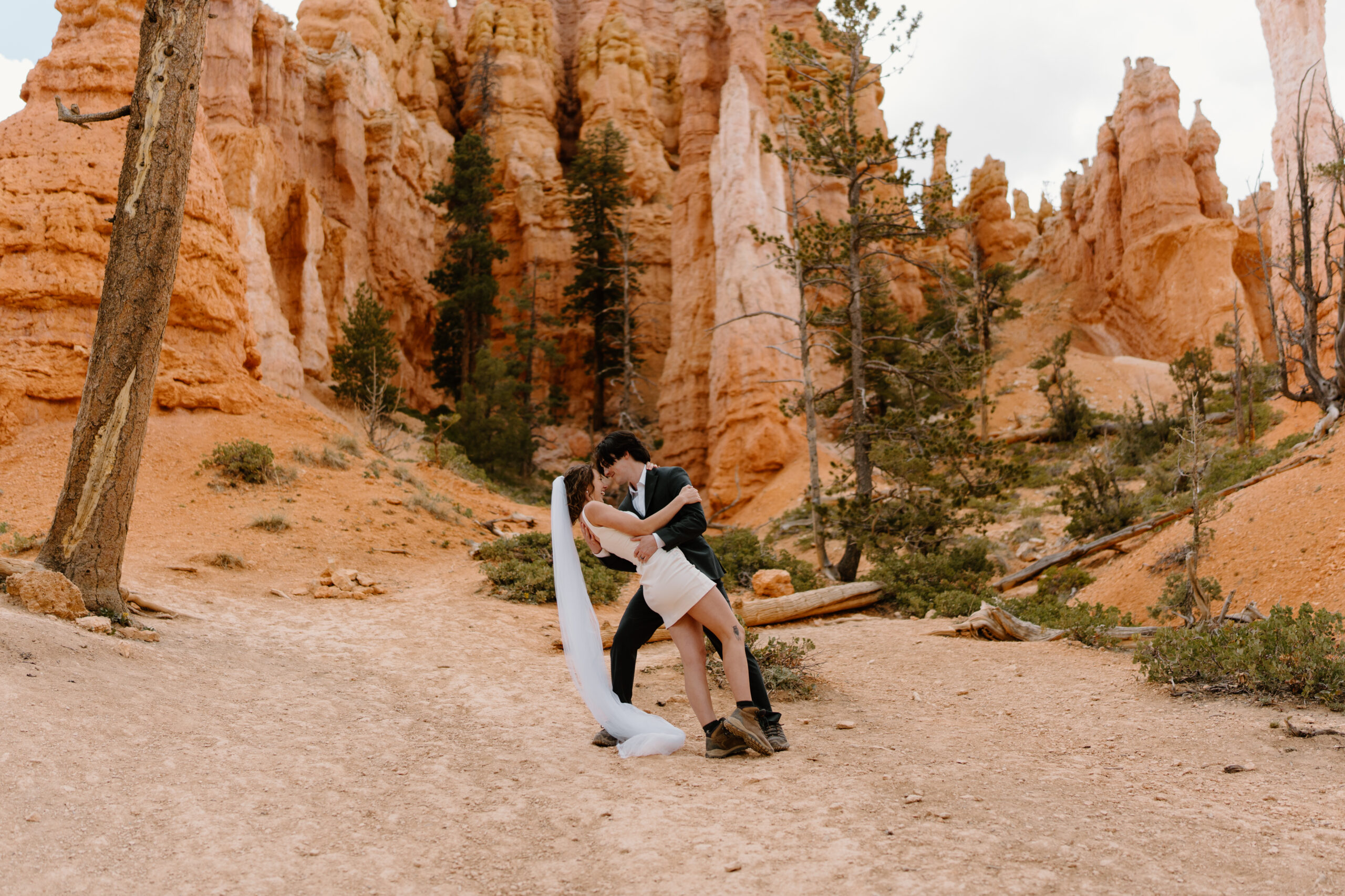 groom (charcoal gray suit, hiking boots) dipping bride (short white dress, long veil, hiking boots) during their non-traditional elopement at Bryce Canyon National Park while on a hike 