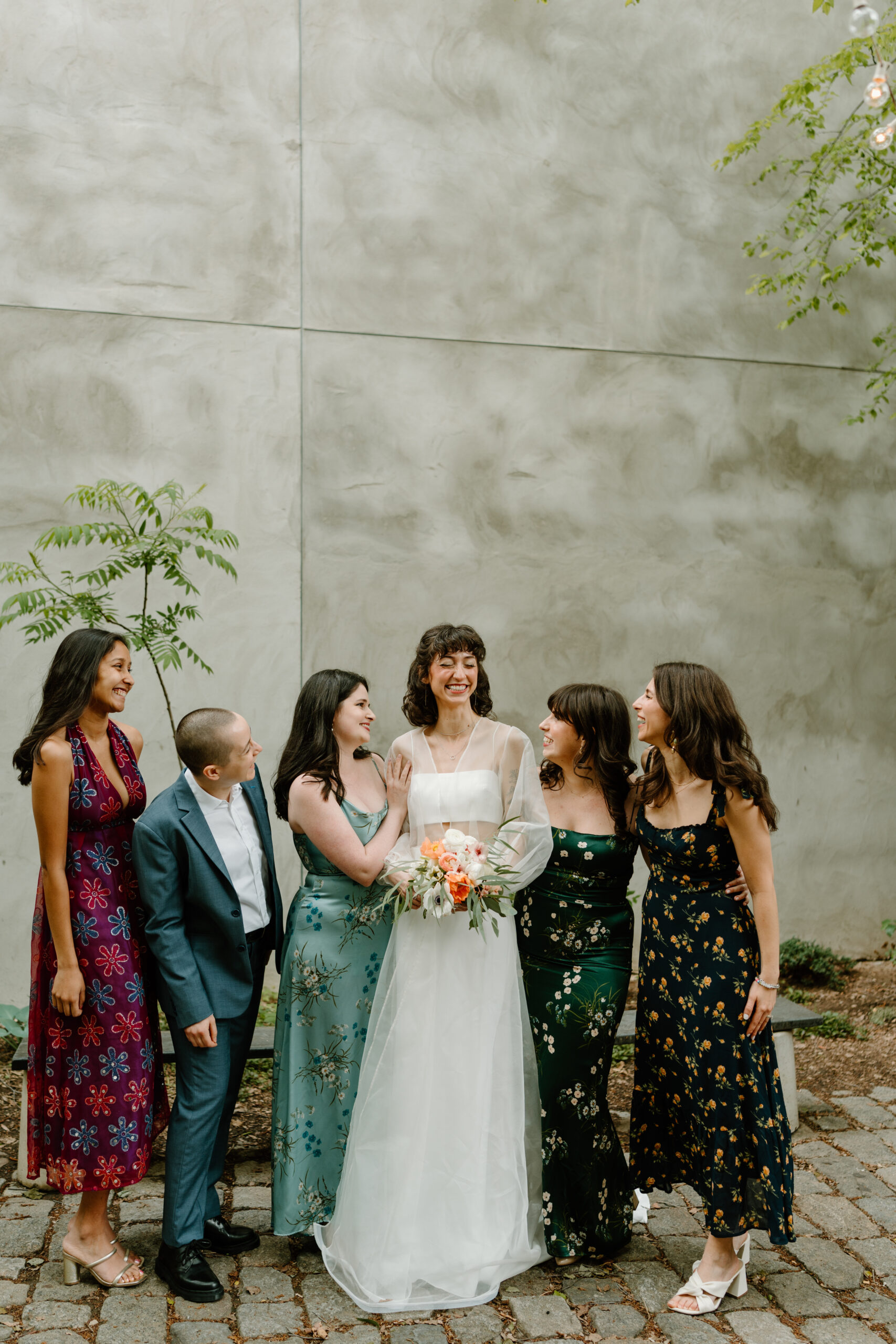 a bride (wearing white) and five friends (four in non-traditional colorful floral dresses, one in a blue suit) smiling at each other