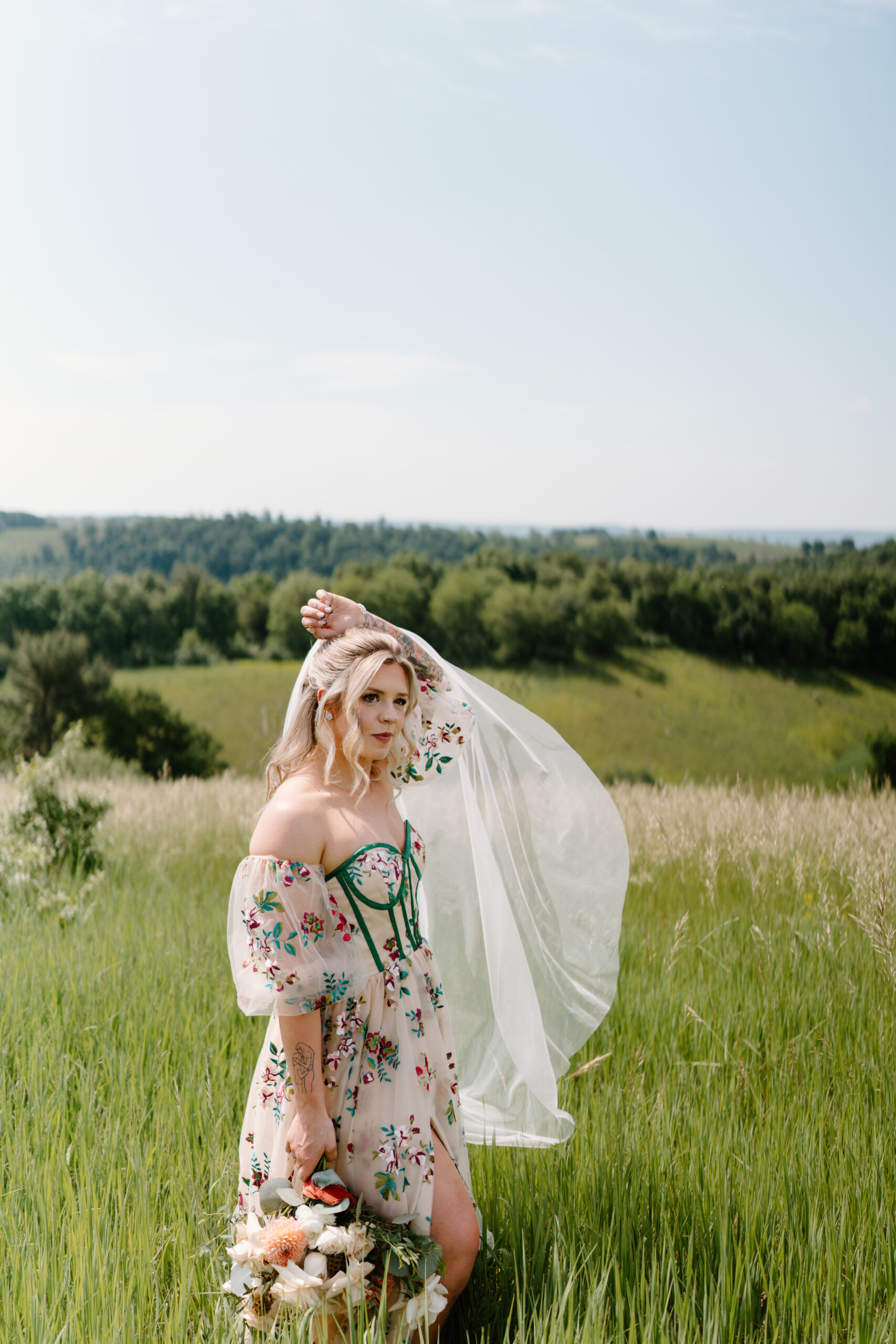 bride with blonde hair tossing her veil in the breeze and holding her tattooed left arm over her head, smiling softly off camera and holding her bouquet in her right hand at her side. her non-traditional wedding dress is pale pink with colorful embroidered flowers and green corset boning.