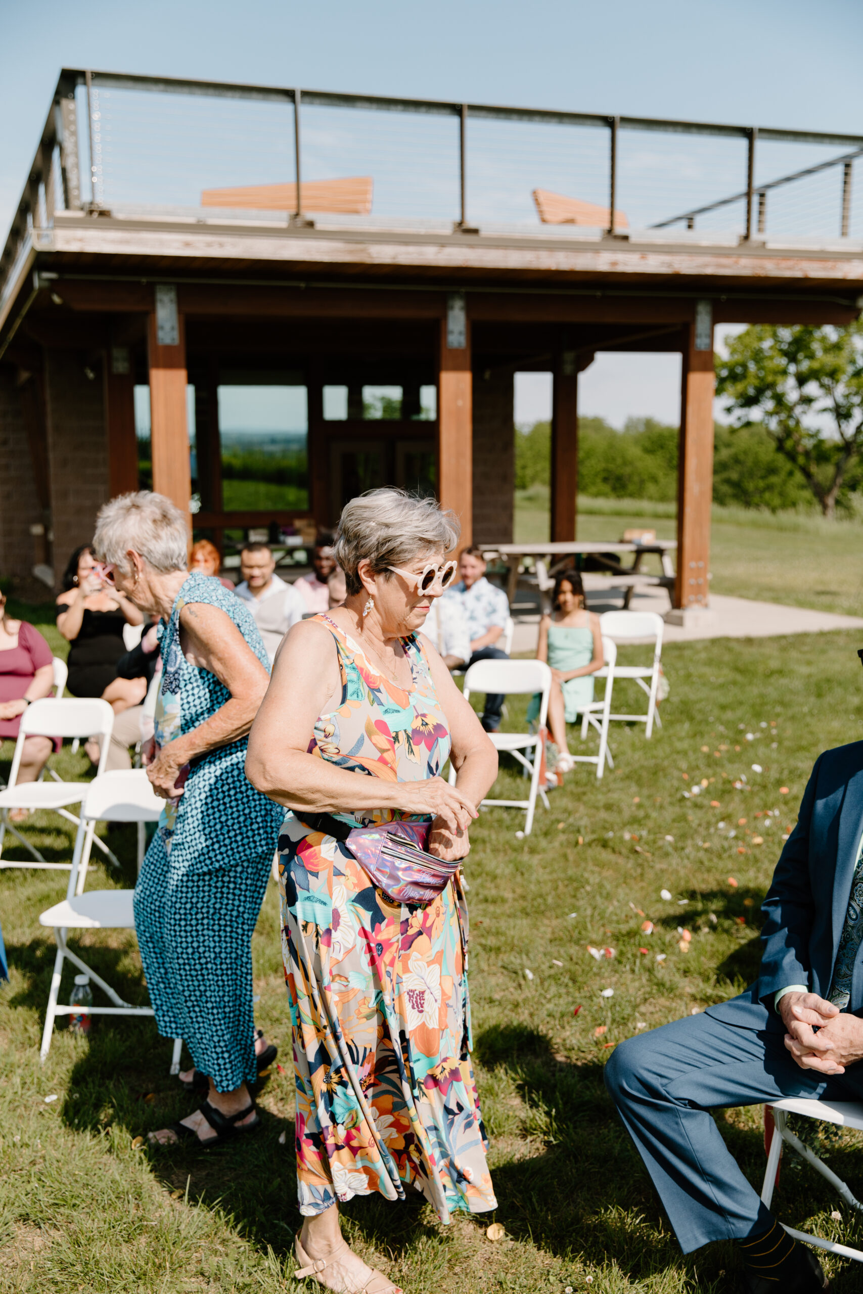 two grandmothers in colorful dresses, serving as the flower girls for a non-traditional wedding ceremony, wearing sunglasses and throwing flower petals out of the fanny packs they wear