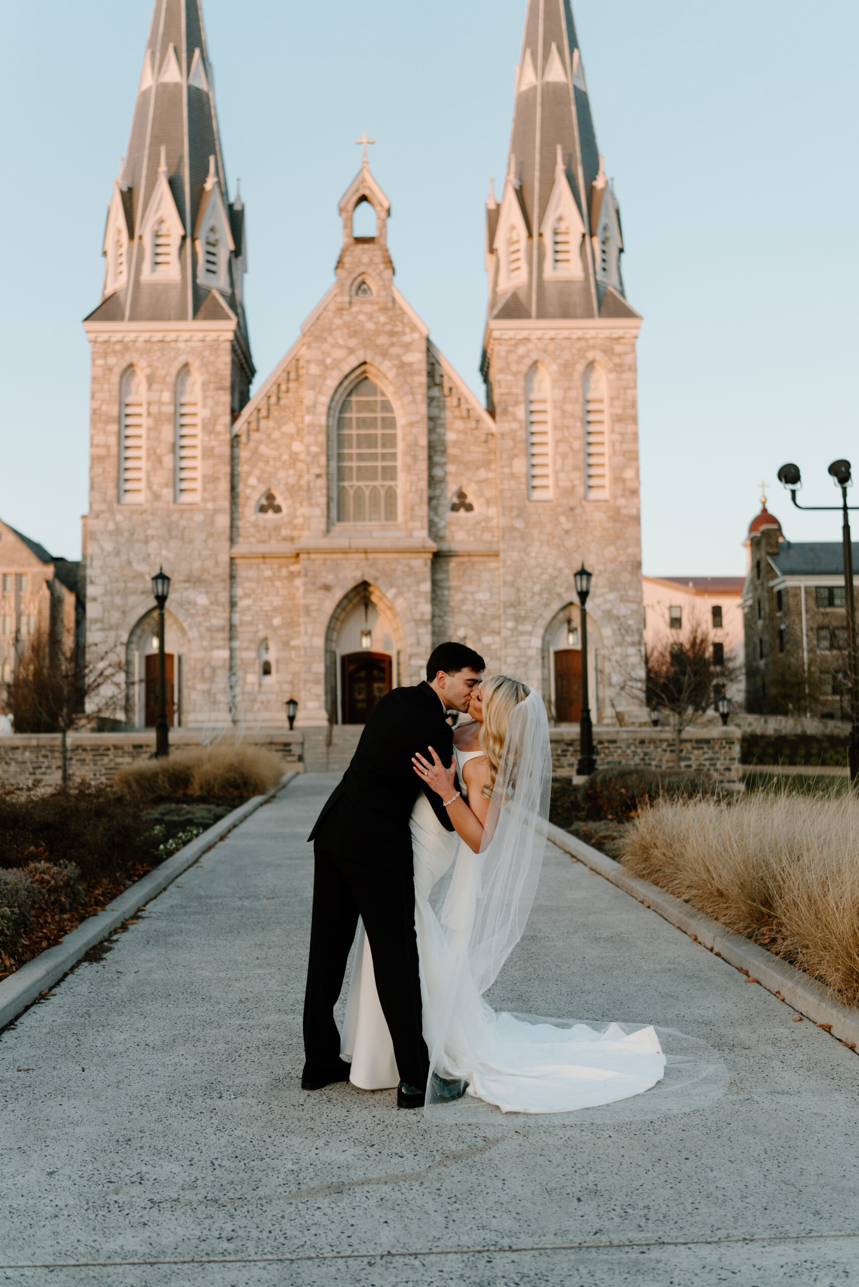 Groom (black suit) dipping and kissing his bride (white dress, cathedral veil) in front of Villanova cathedral after their wedding ceremony