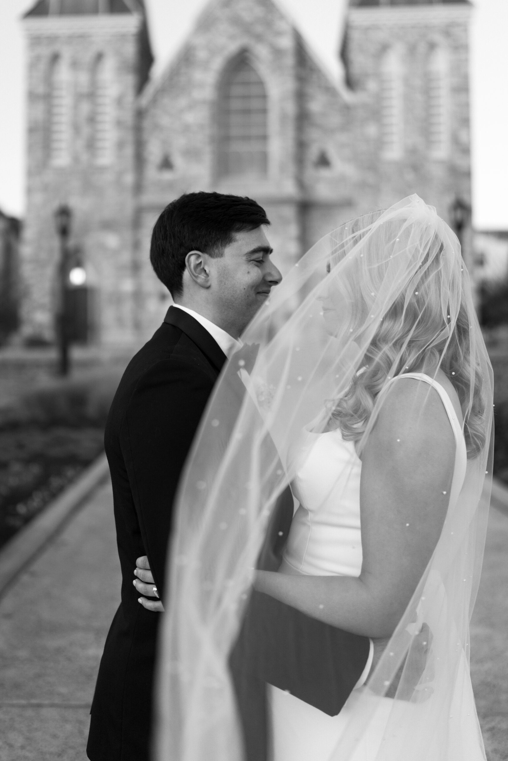black and white image of groom (black suit) and bride (blonde hair, white dress) embracing and smiling at each other while her veil blows in front of them on their wedding day