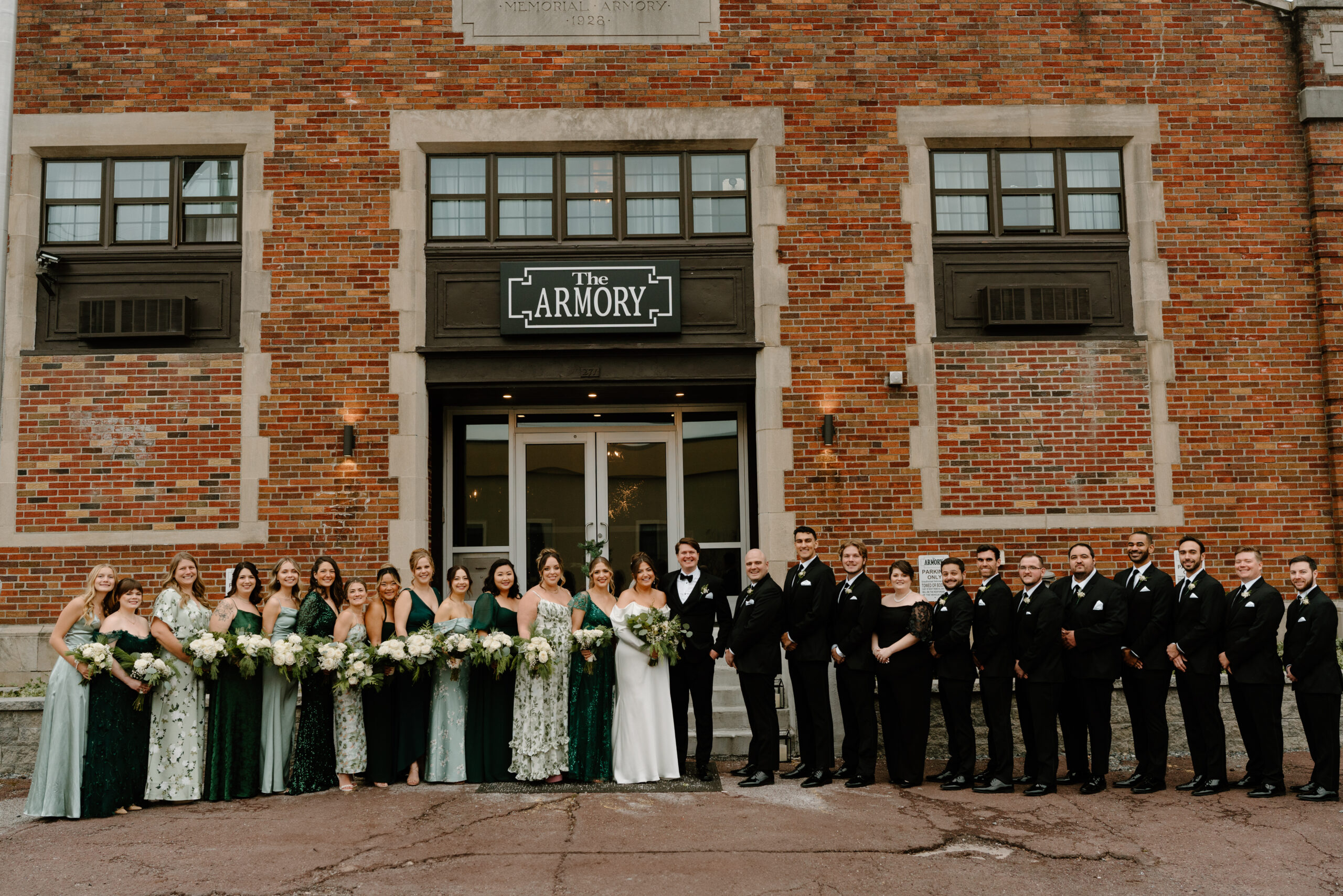 a large non-traditional wedding party portrait of mixed genders, the bride's side wearing green patterned dresses and the groom's side wearing all black dresses and suits. All are smiling at the camera in front of a brick building.
