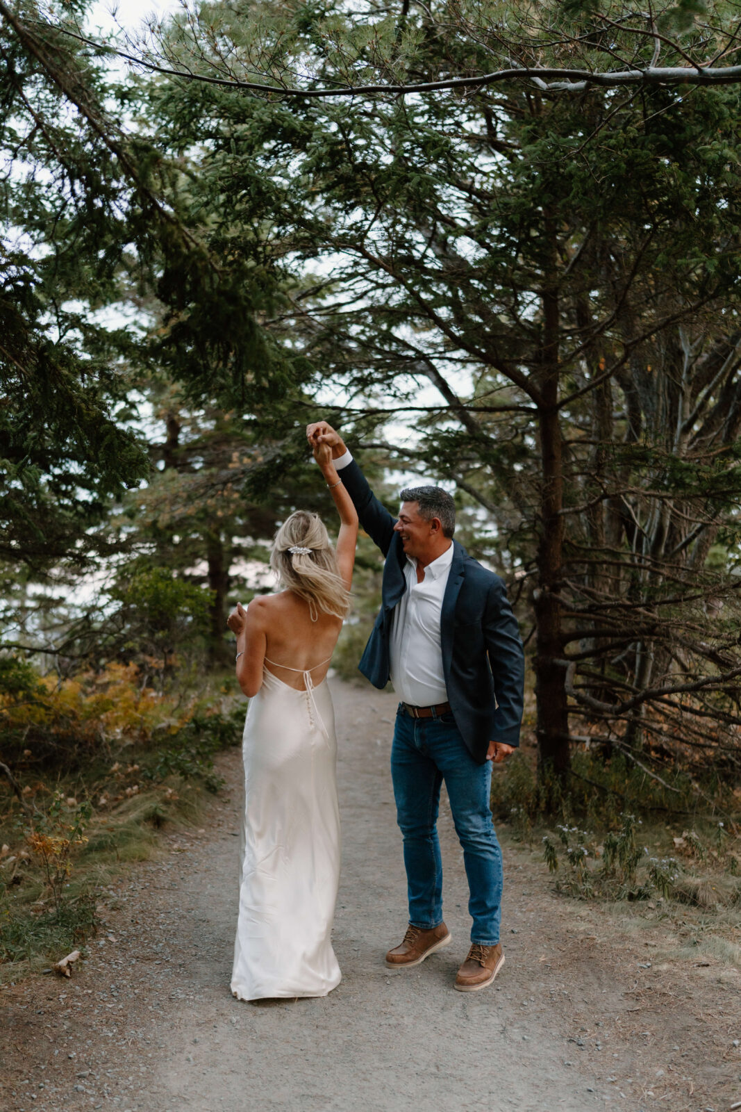  a man (jeans, white shirt, navy blue blazer) twirling his wife (blonde hair, white backless sleek wedding dress) and smiling, on a dirt trail surrounded by evergreen trees after eloping in pennsylvania