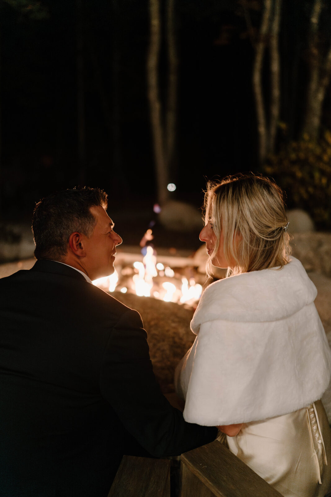close up image of a bride (blonde, in a white dress and a furry white shaw) and groom (salt and pepper hair, dark blue suit jacket) smiling at each other while sitting in front of a bonfire after eloping