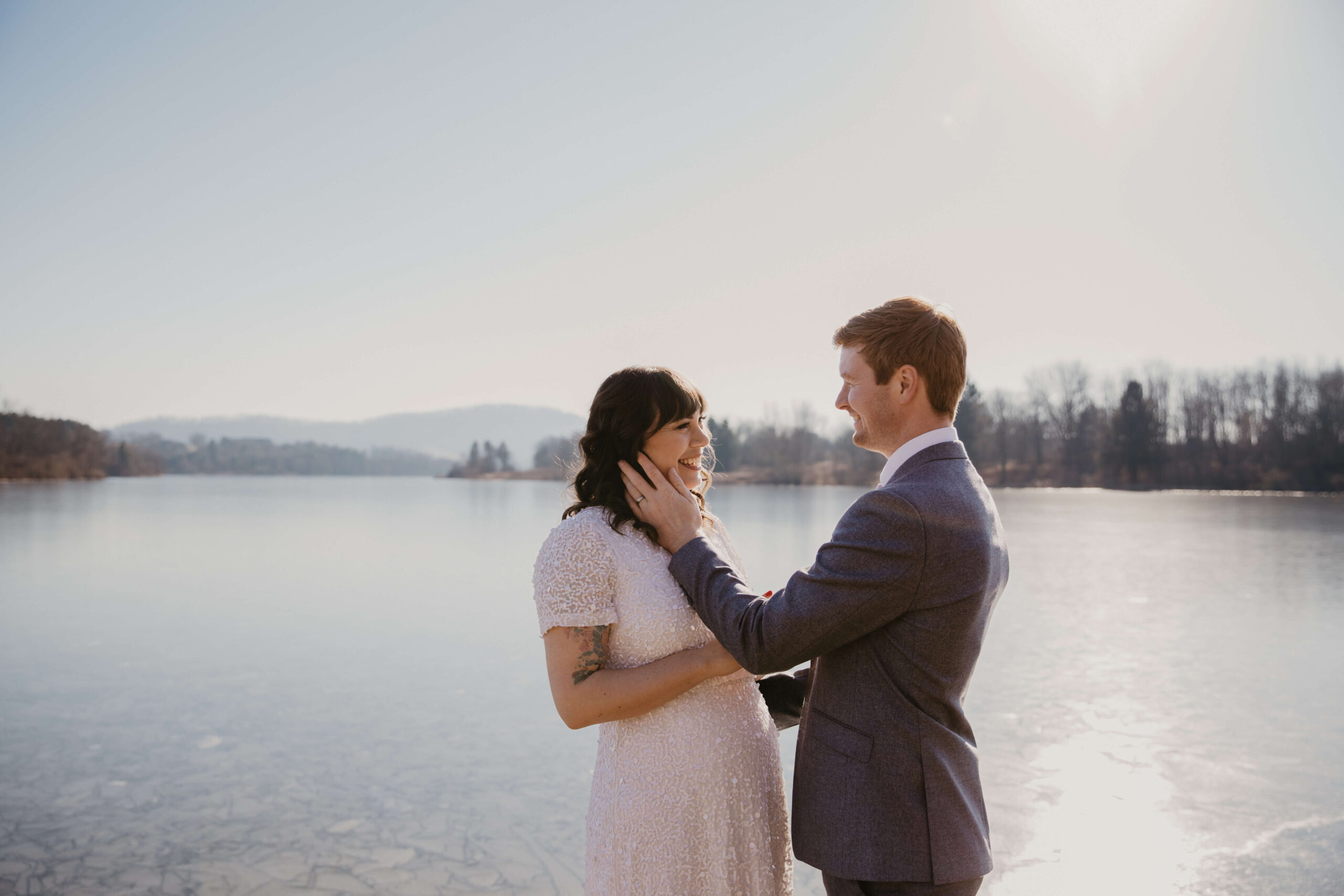 bride (brown hair with bangs and a sequined white dress) smiling at her groom (gray suit, red hair) who is holding her face with his left hand, about to kiss her. They are standing in front of a frozen lake in bright sun, with a mountain in the distance, after eloping in Pennsylvania