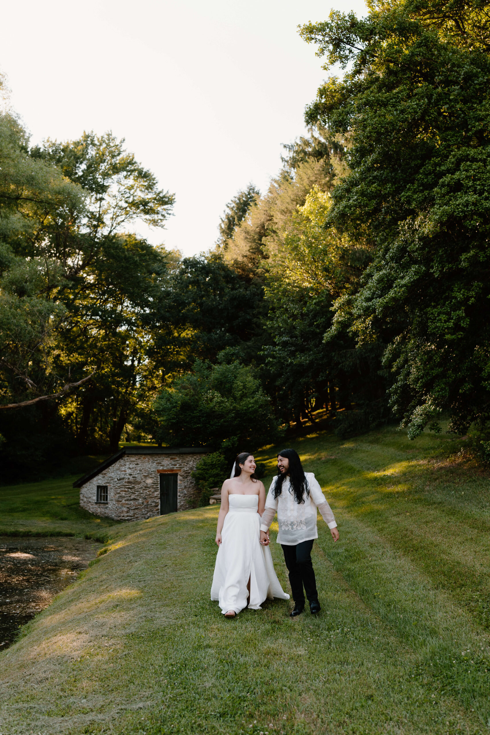 a bride and groom (both long brown hair) walking toward the camera in a green grassy field surrounded by tall trees, after eloping in the Pennsylvania countryside