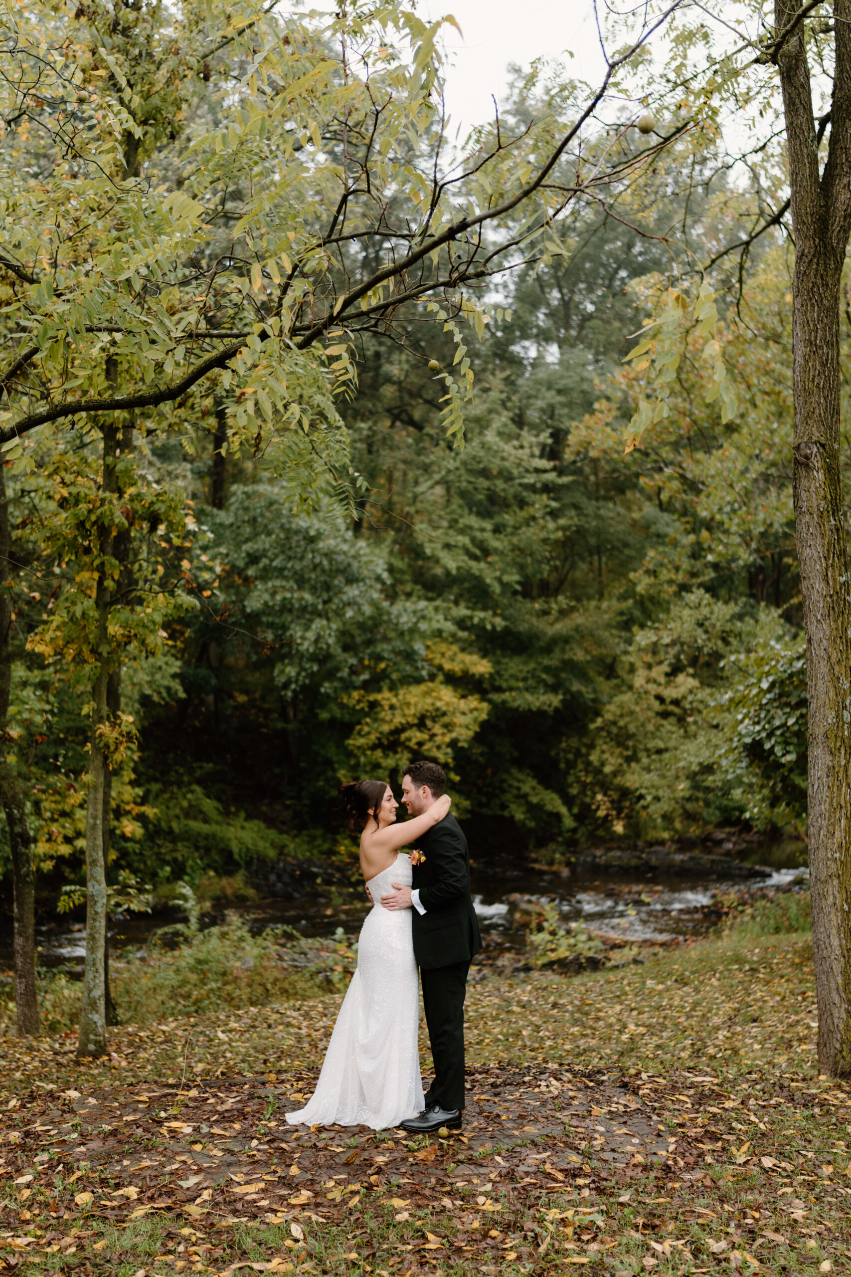 bride (brown hair in an updo) and groom (brown hair, black suit and a boutonniere) embracing in front of a river, surrounded by fall leaves on a rainy Pennsylvania day after eloping