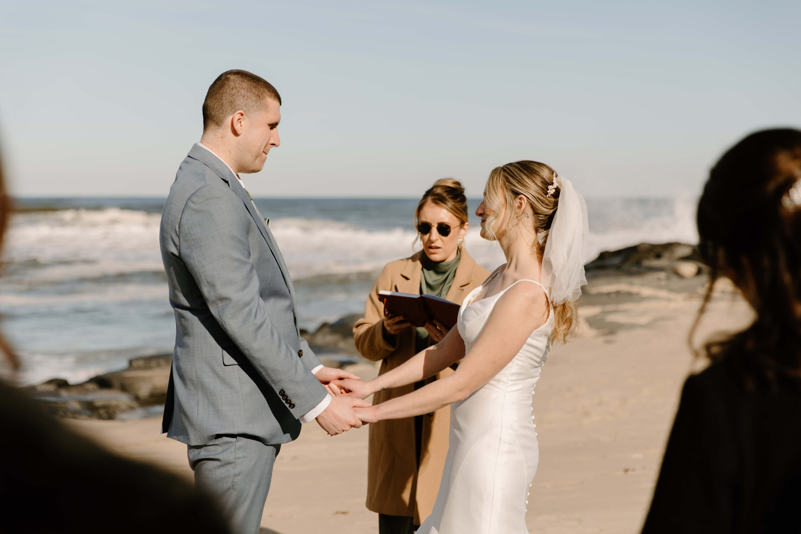 bride and groom holding hands during their elopement ceremony at the beach, while their officiant performs the ceremony wearing a jacket. The ocean waves crash behind them on the rocks while they elope