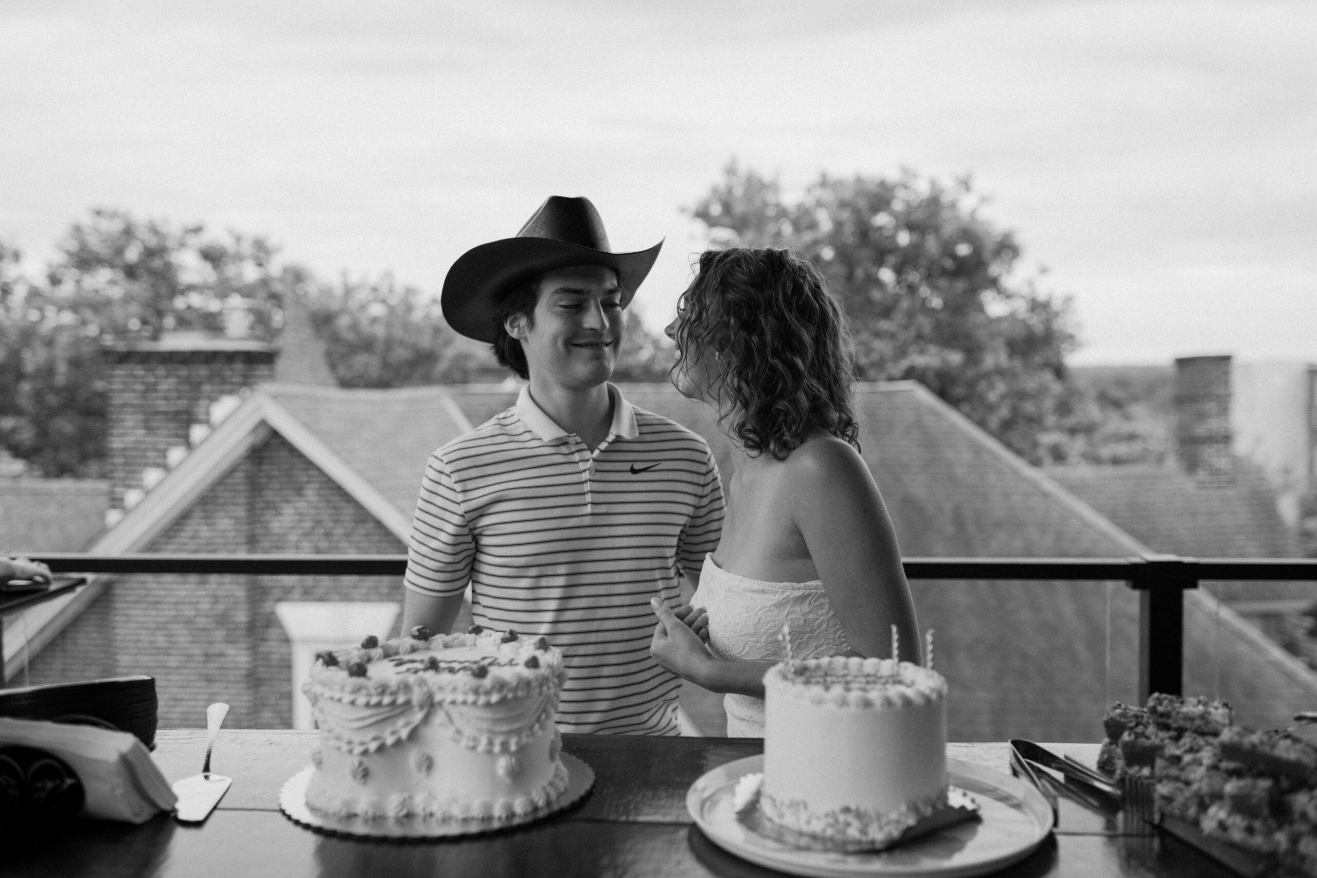 black and white image of a bride (strapless white dress) and groom (striped polo shirt, cowboy hat) smiling at each other while standing over their vintage decorated wedding cake during an elopement celebration