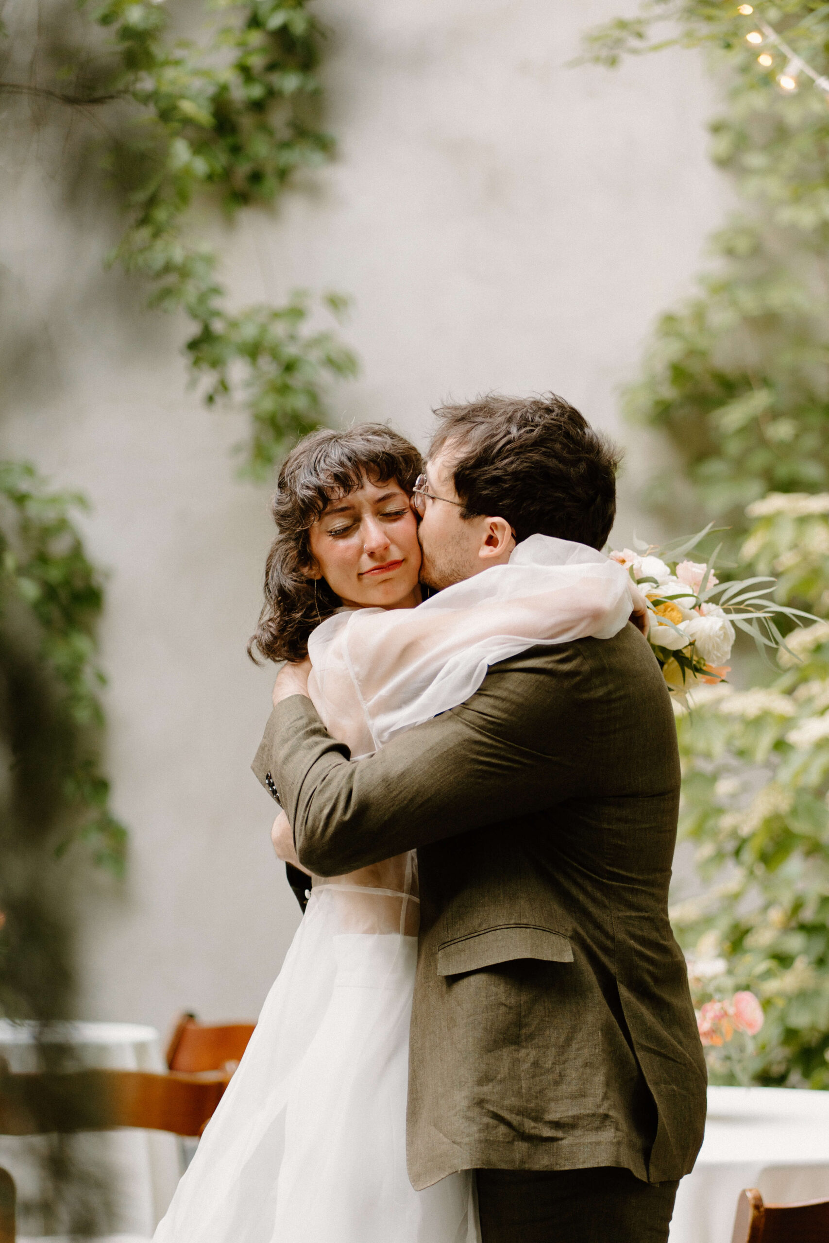 a groom (brown hair, olive green suit jacket) hugging and kissing his bride (sheer white dress) on the cheek as she closes her eyes and holds back tears after their pennsylvania elopement