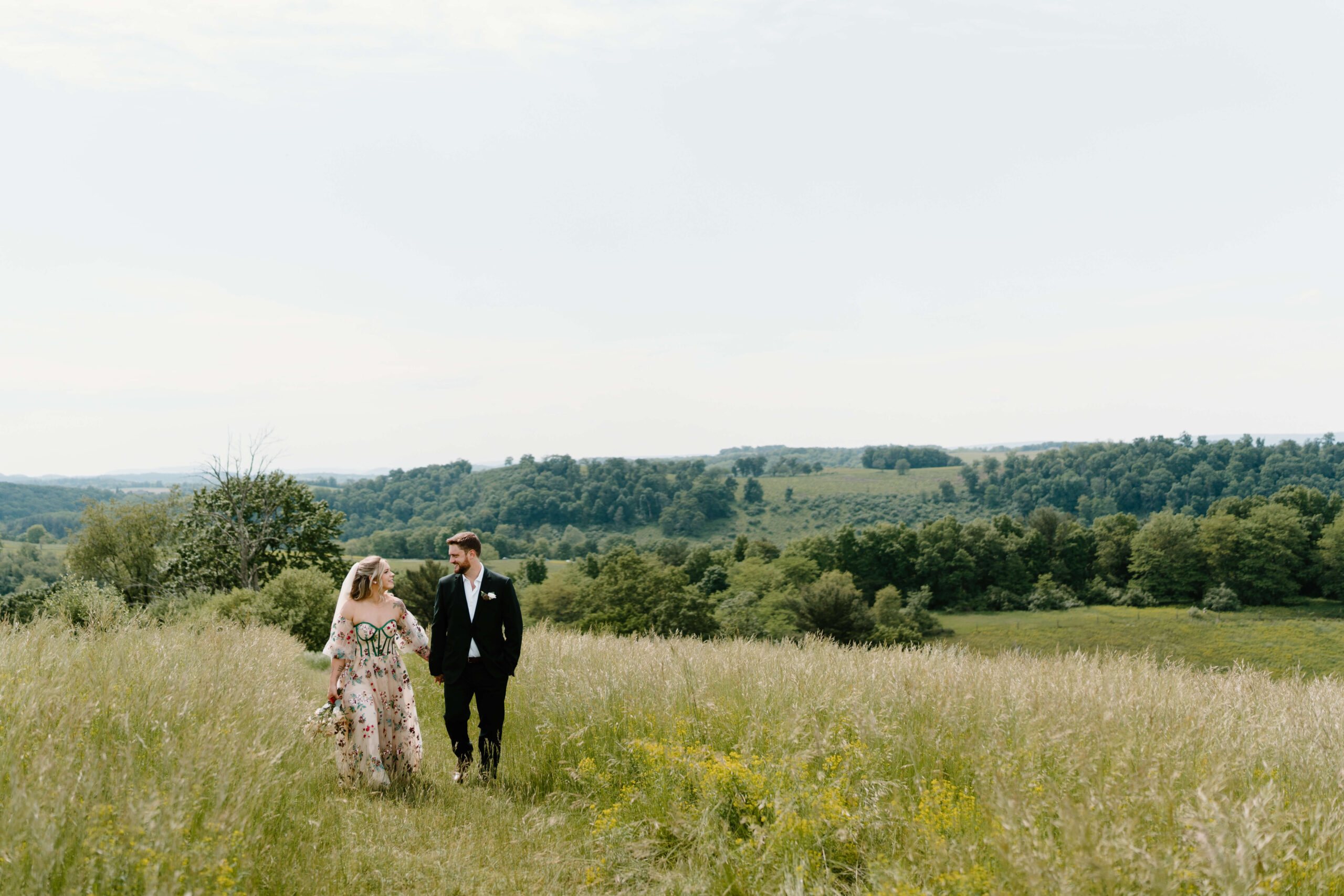 bride (blonde hair and a pink wedding gown embroidered with emerald green boning and bright, colorful florals) and groom (dark green suit, brown hair) smiling at each other while holding hands and walking toward the camera through a field of tall grass, rolling hills behind them, on their elopement day