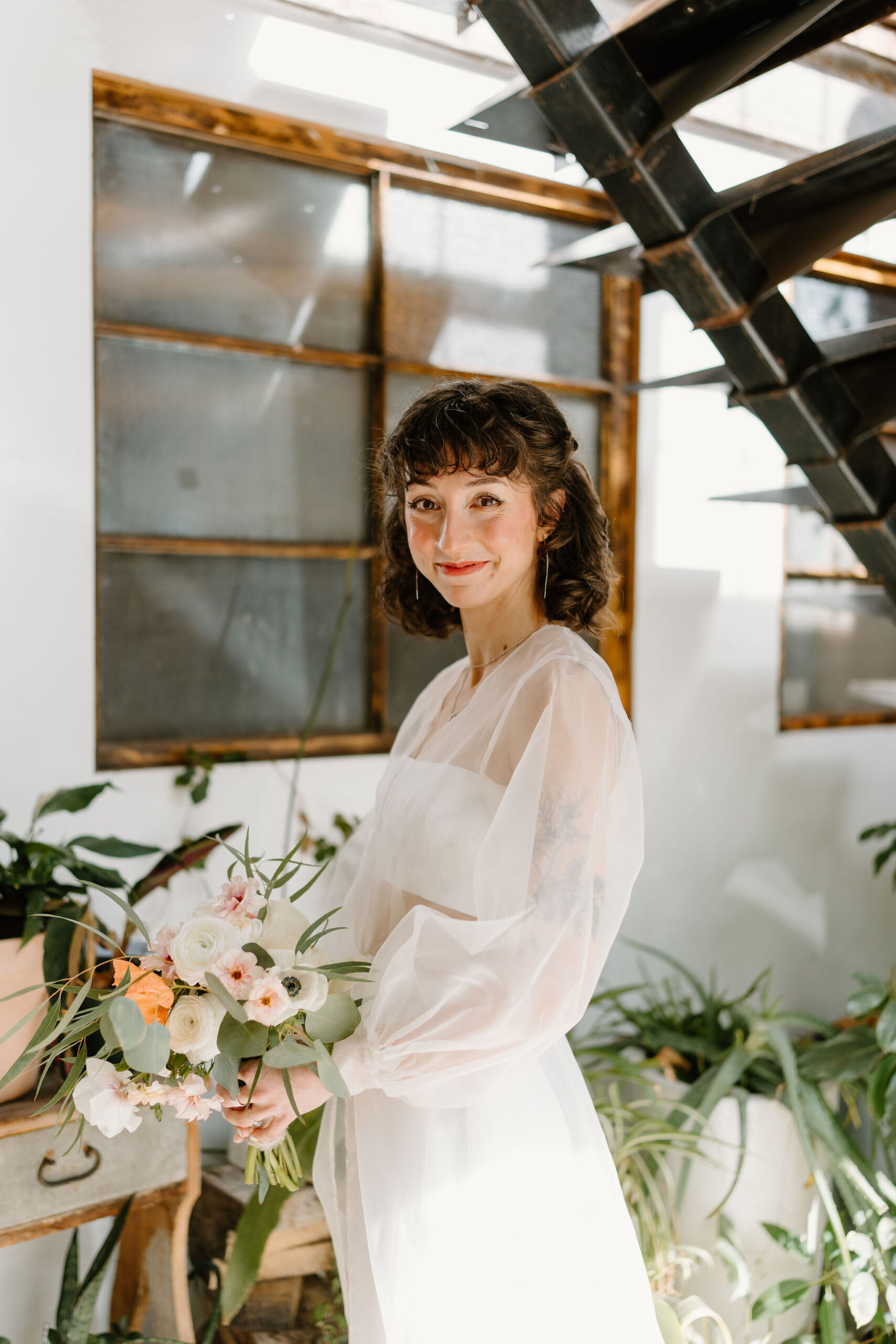 bride (short brown curly hair, bangs) holding her bouquet and smiling softly toward the camera in front of a wall full of plants and an industrial staircase at the Maas Building