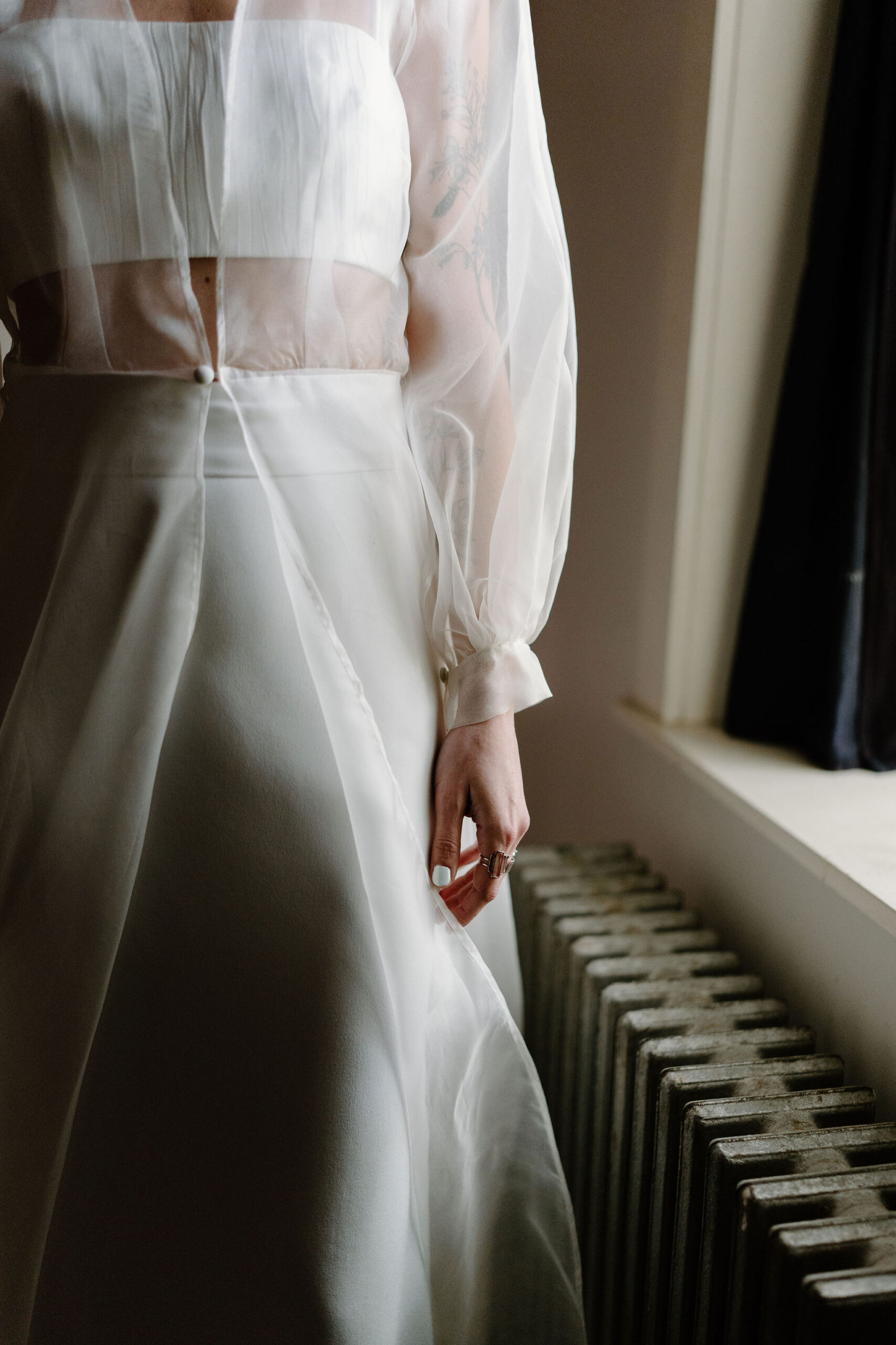 close up image of a bride wearing a two piece cropped spaghetti strap top, a simple skirt, and a sheer cape with one button at her waist, her hand held at her side with lots of rings at the Maas Building